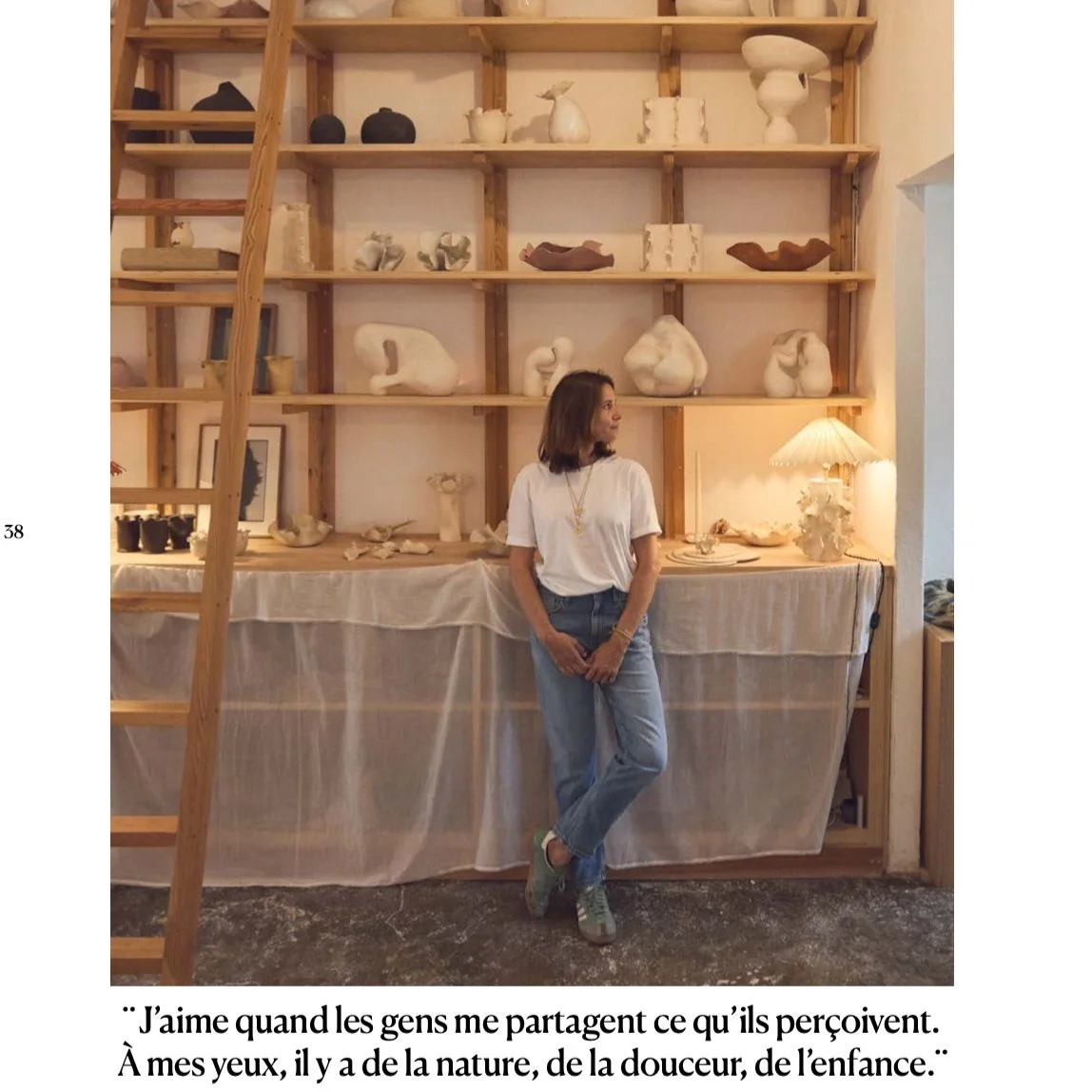 A woman standing in front of a wooden shelf filled with various white ceramic sculptures and decorative items. She is wearing a white T-shirt, jeans, and sneakers, and is looking to her right. There is a table covered with a cloth and a small lamp to
