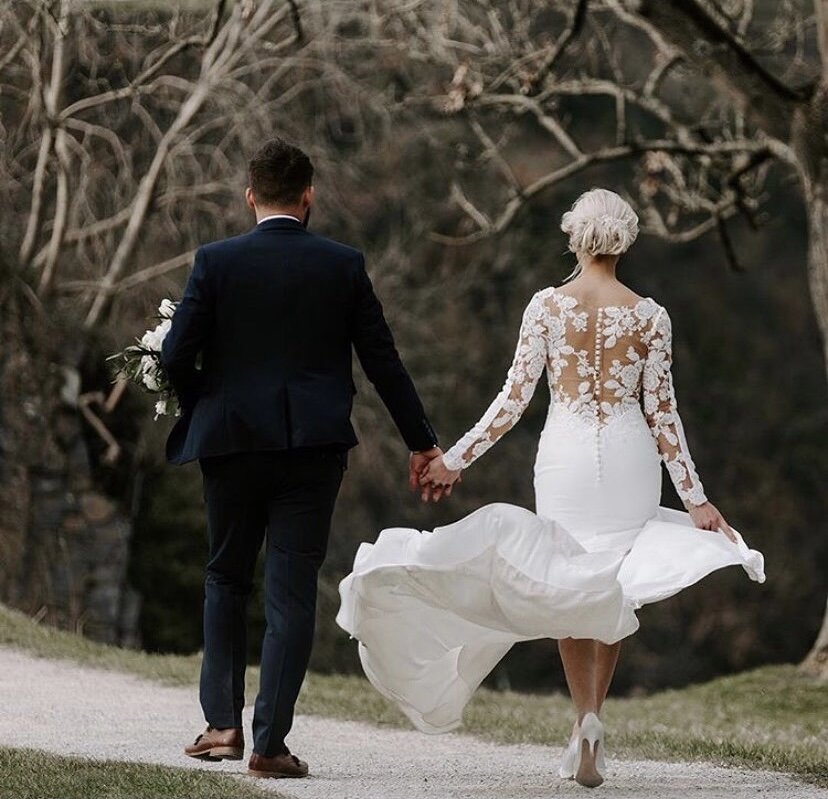 Bride and groom holding hands walking outdoors, bride in white lace dress, groom in dark suit, trees in background.