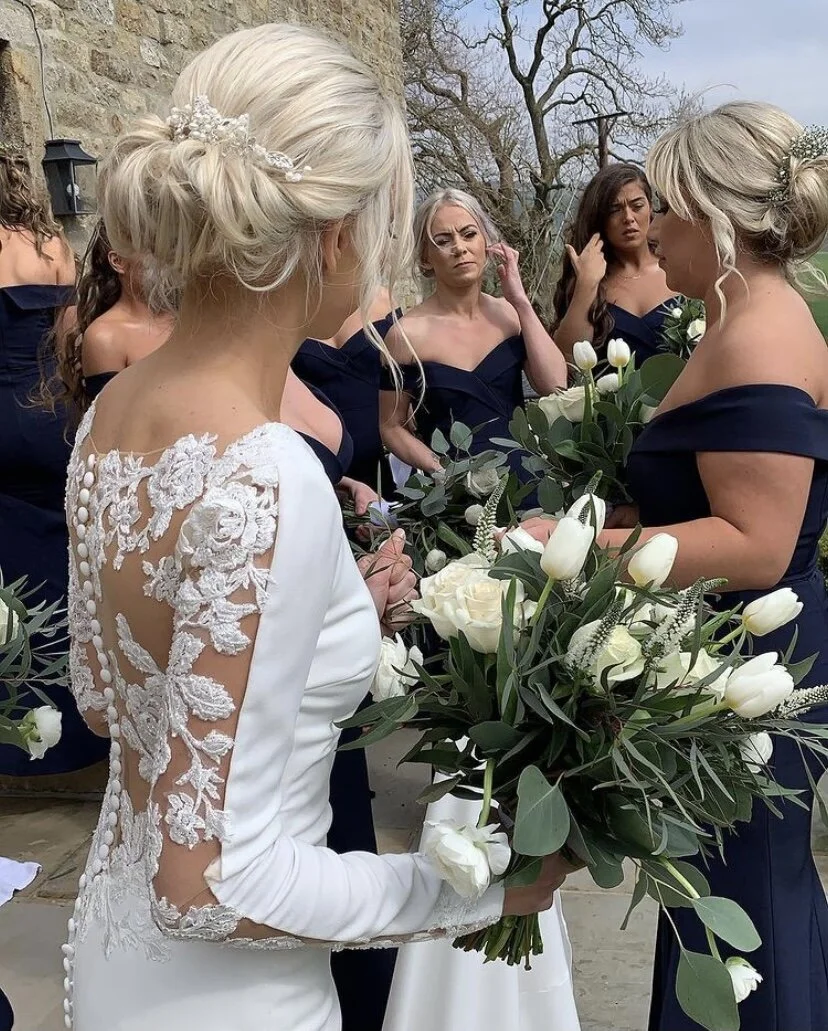 A bride in a white wedding dress with lace sleeves holding a bouquet of white roses and tulips, surrounded by bridesmaids in dark blue dresses during a wedding ceremony outdoors.