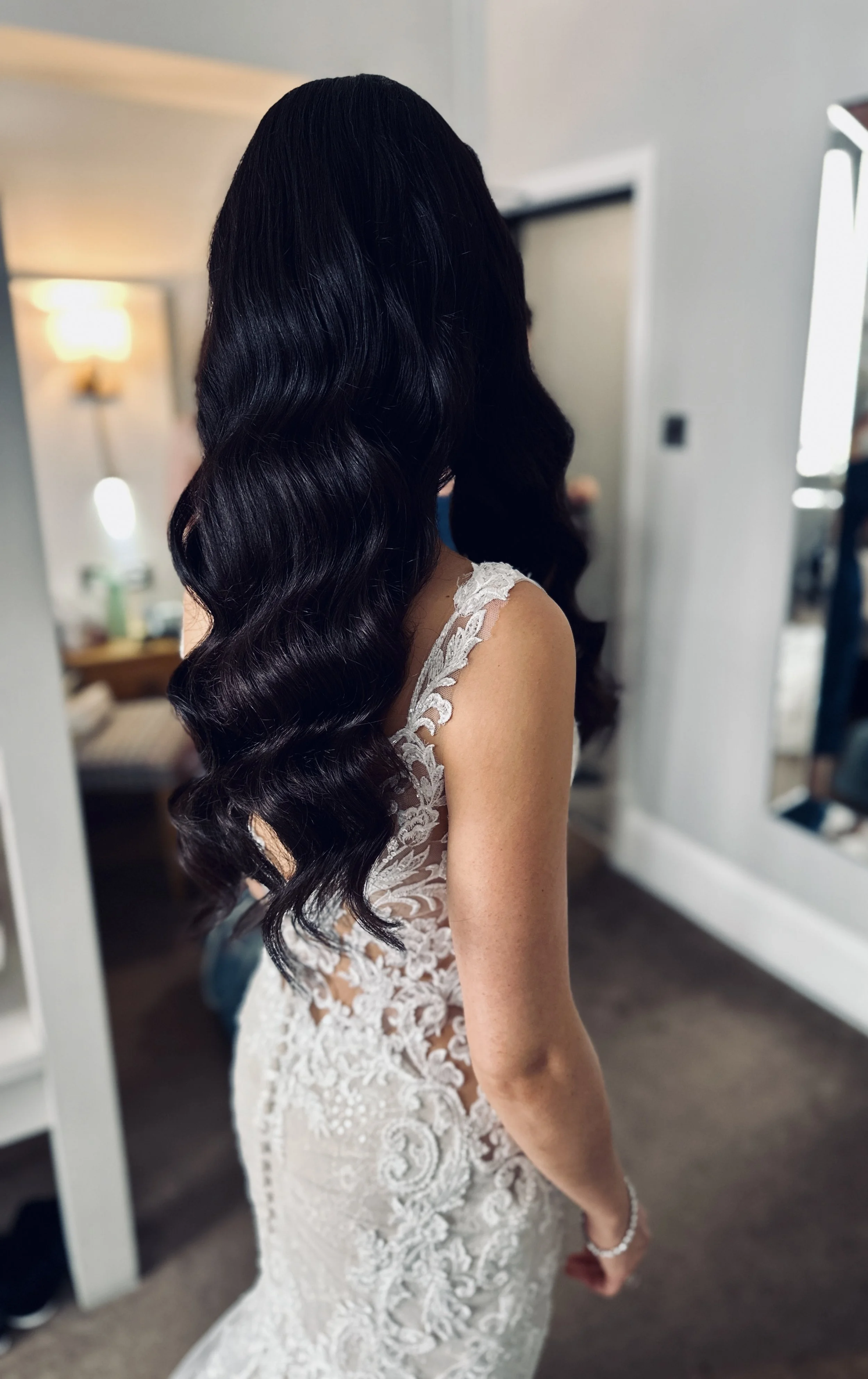 A woman with long, wavy black hair wearing a lace wedding dress with sheer lace details on the back, standing in a room with a mirror and a lamp in the background.