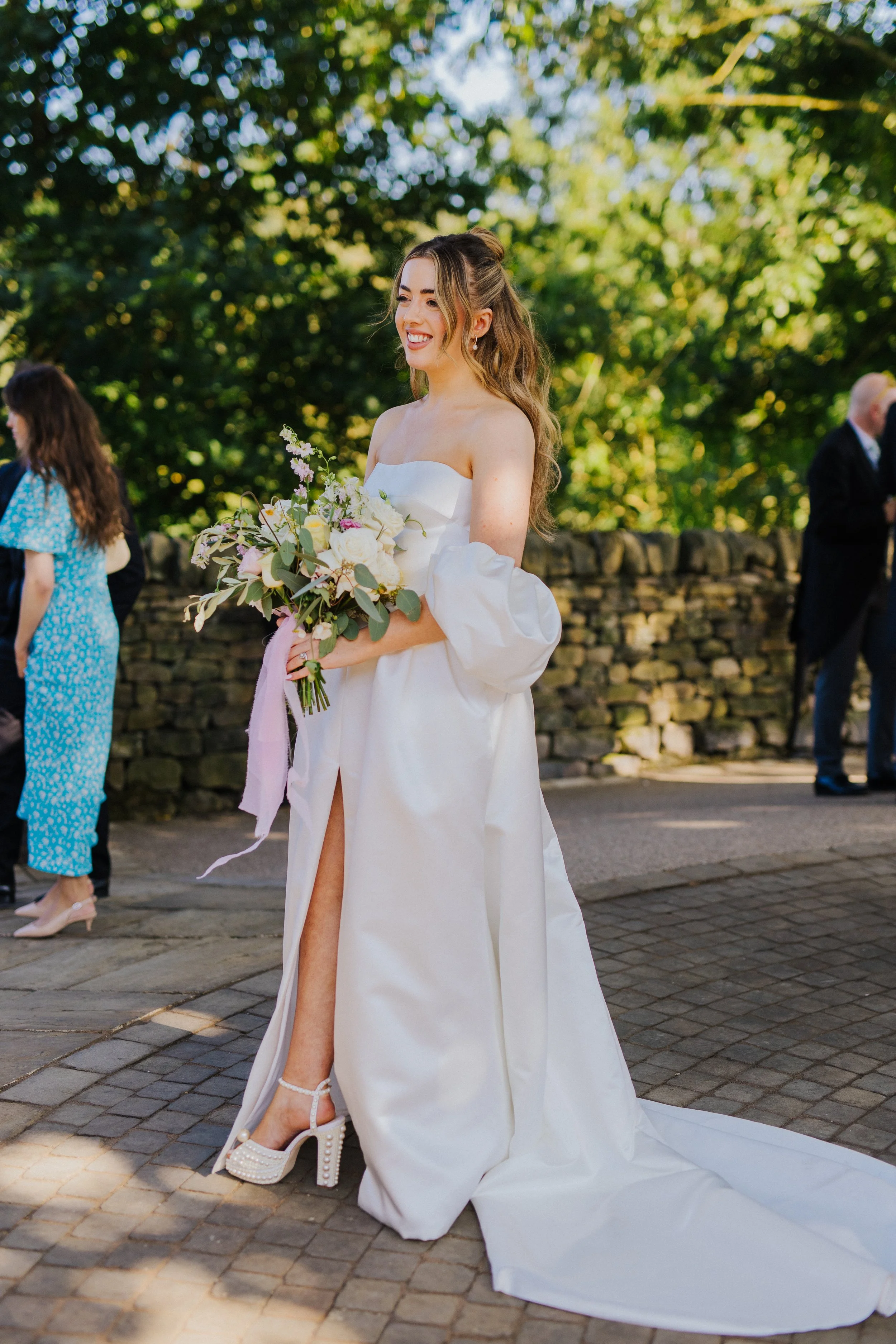 A smiling bride in a white wedding dress holding a bouquet of flowers at an outdoor wedding.