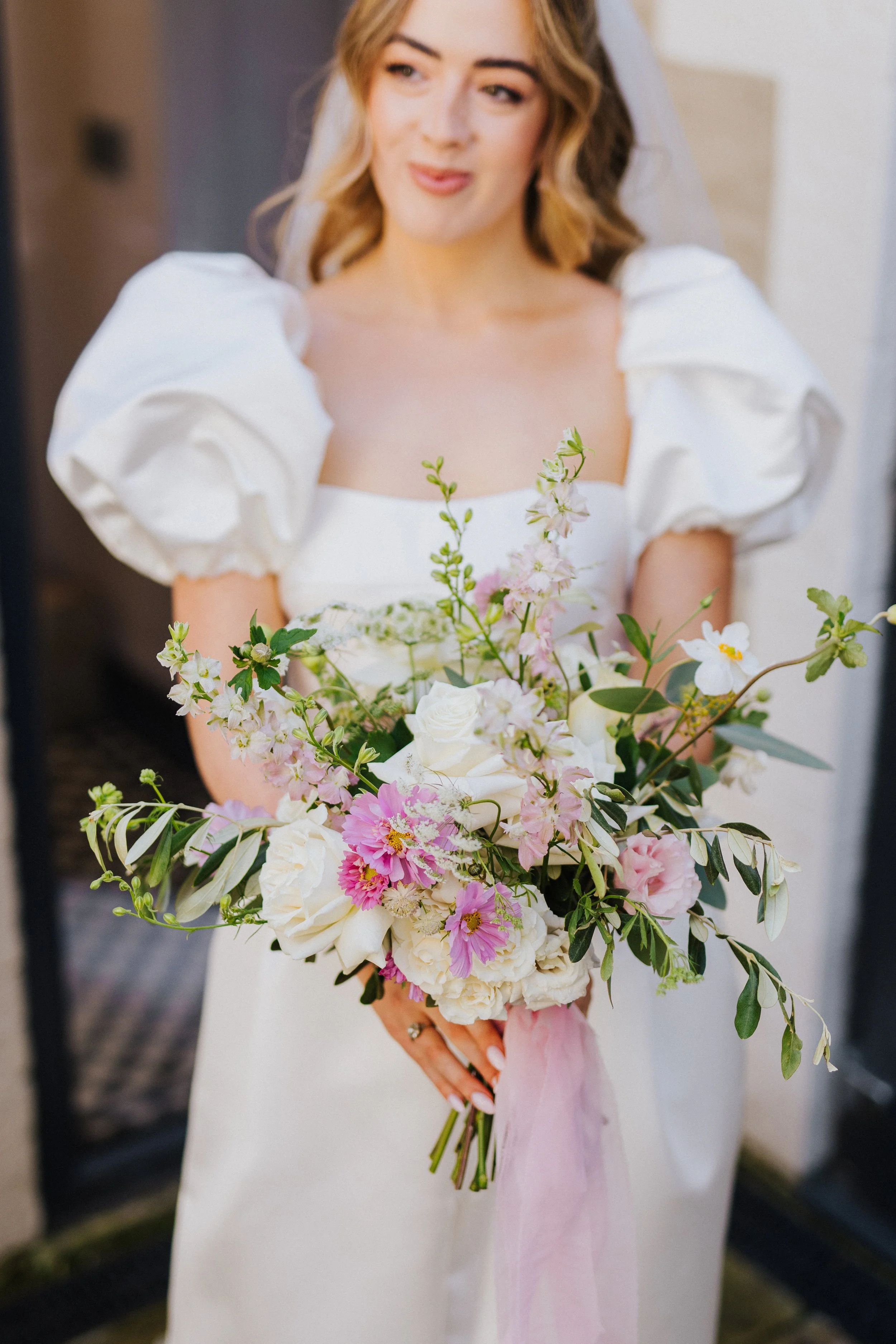 A woman in a white dress holding a bouquet of white and pink flowers. She has blonde hair and is looking slightly to the side with a slight smile.