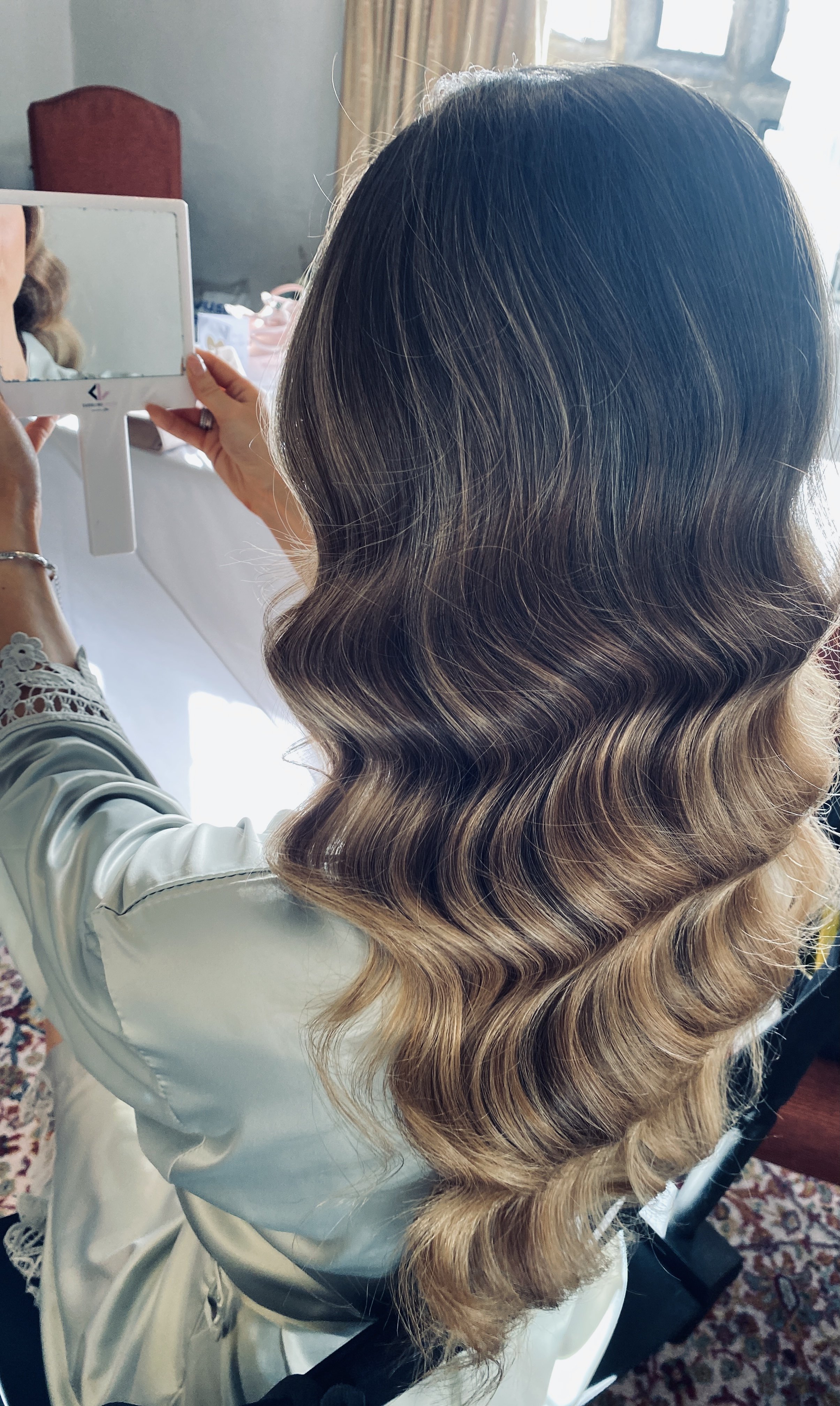 A woman with long, wavy, ombre hair in shades of dark blonde and light brown, sitting at a vanity and holding a mirror to look at her hair.