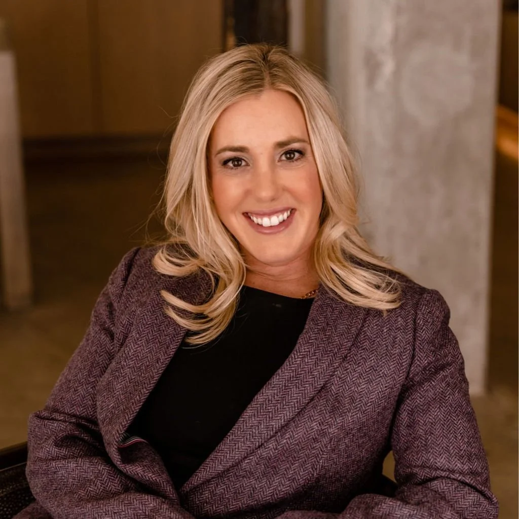 A smiling woman with blonde hair wearing a brown blazer and black top, sitting indoors with a neutral background.