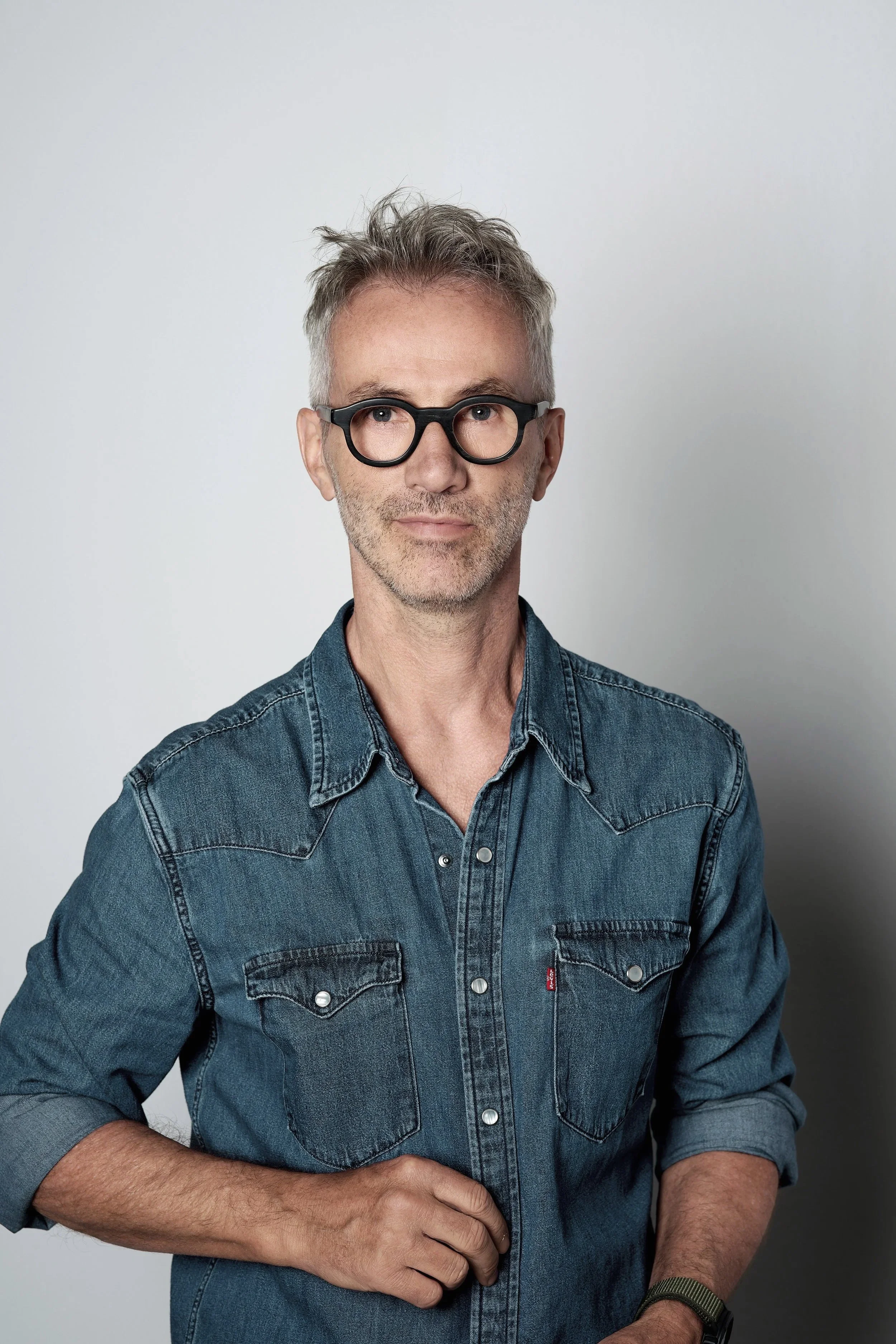 Ein Mann mit grauem, unordentlichem Haar, Brille und einem dunkelblauen Jeanshemd steht vor einem weißen Hintergrund.