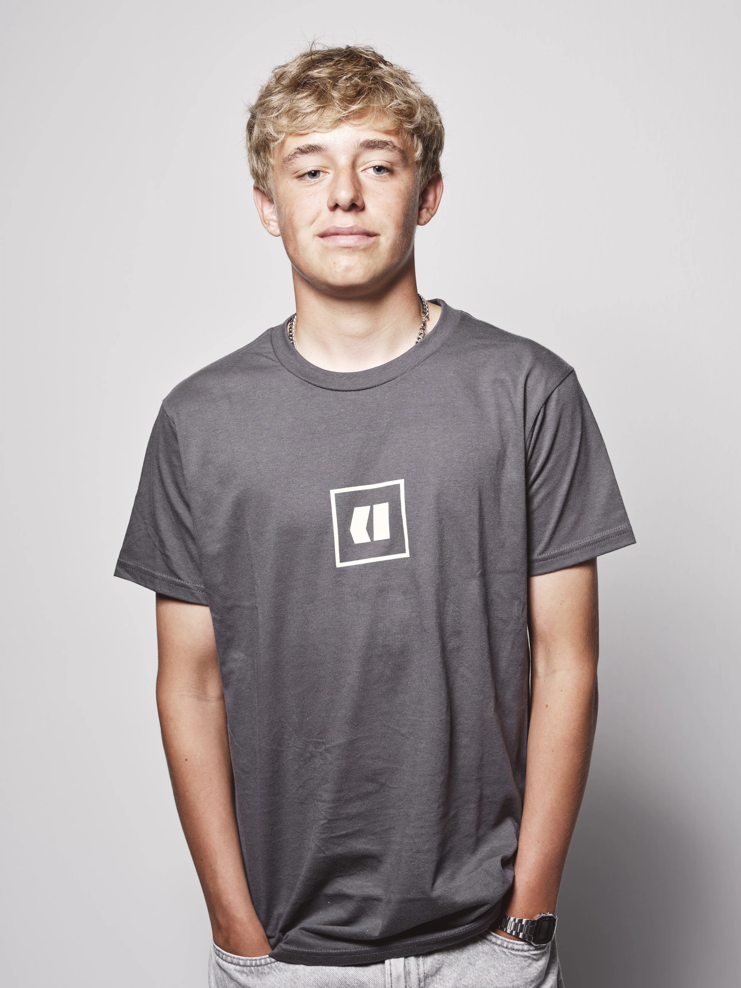 Ein junger Mann mit blondem, lockigem Haar trägt ein T-Shirt mit einem weißen Logo auf der Brust, steht vor einem neutralen Hintergrund.