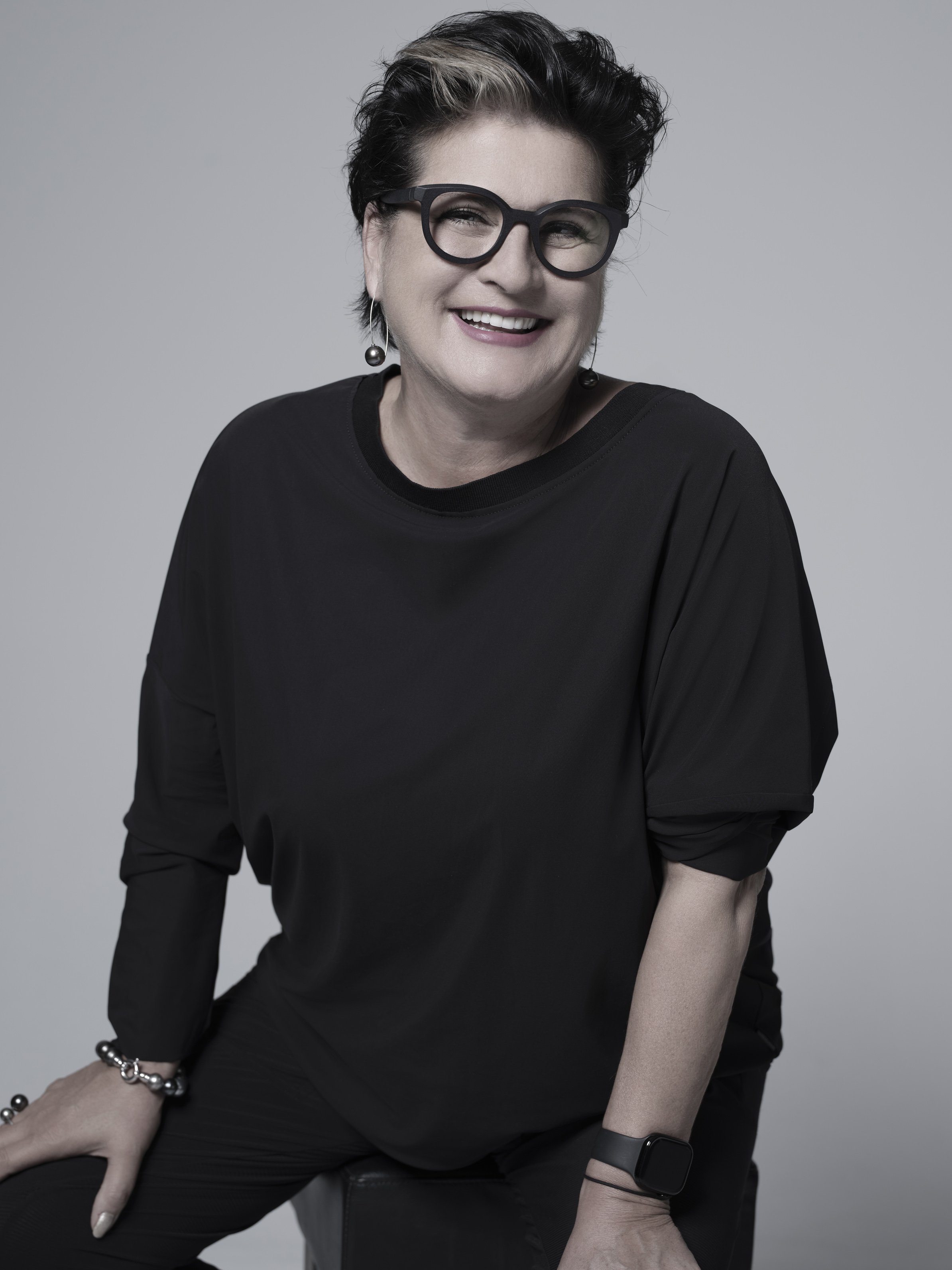 Lächelnde Frau mit schwarzen Haaren, Brille und schwarzer Kleidung sitzt vor grauem Hintergrund.