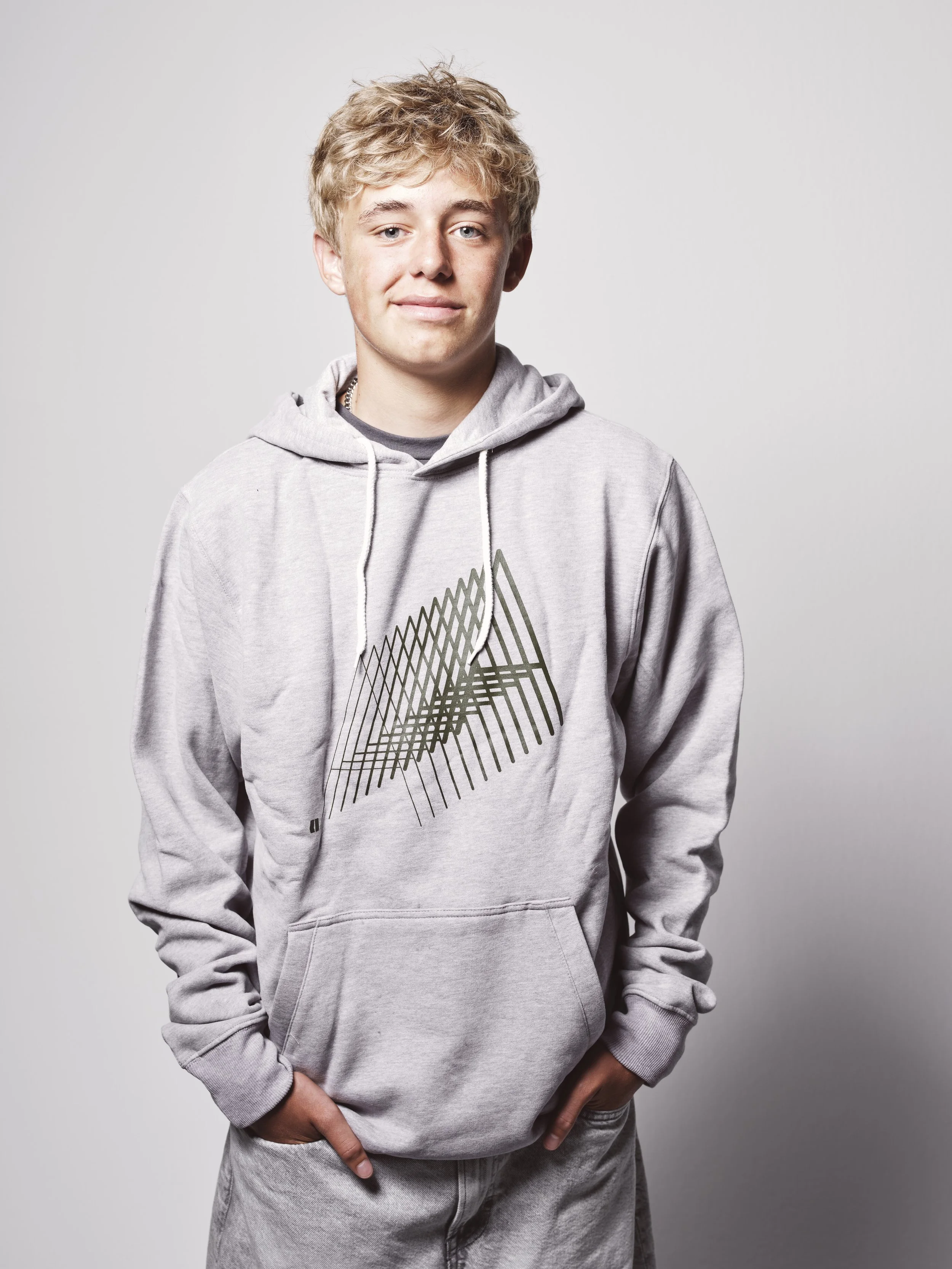 Junge mit blonden, lockigen Haaren trägt grauen Hoodie, steht cool vor hellem Hintergrund.