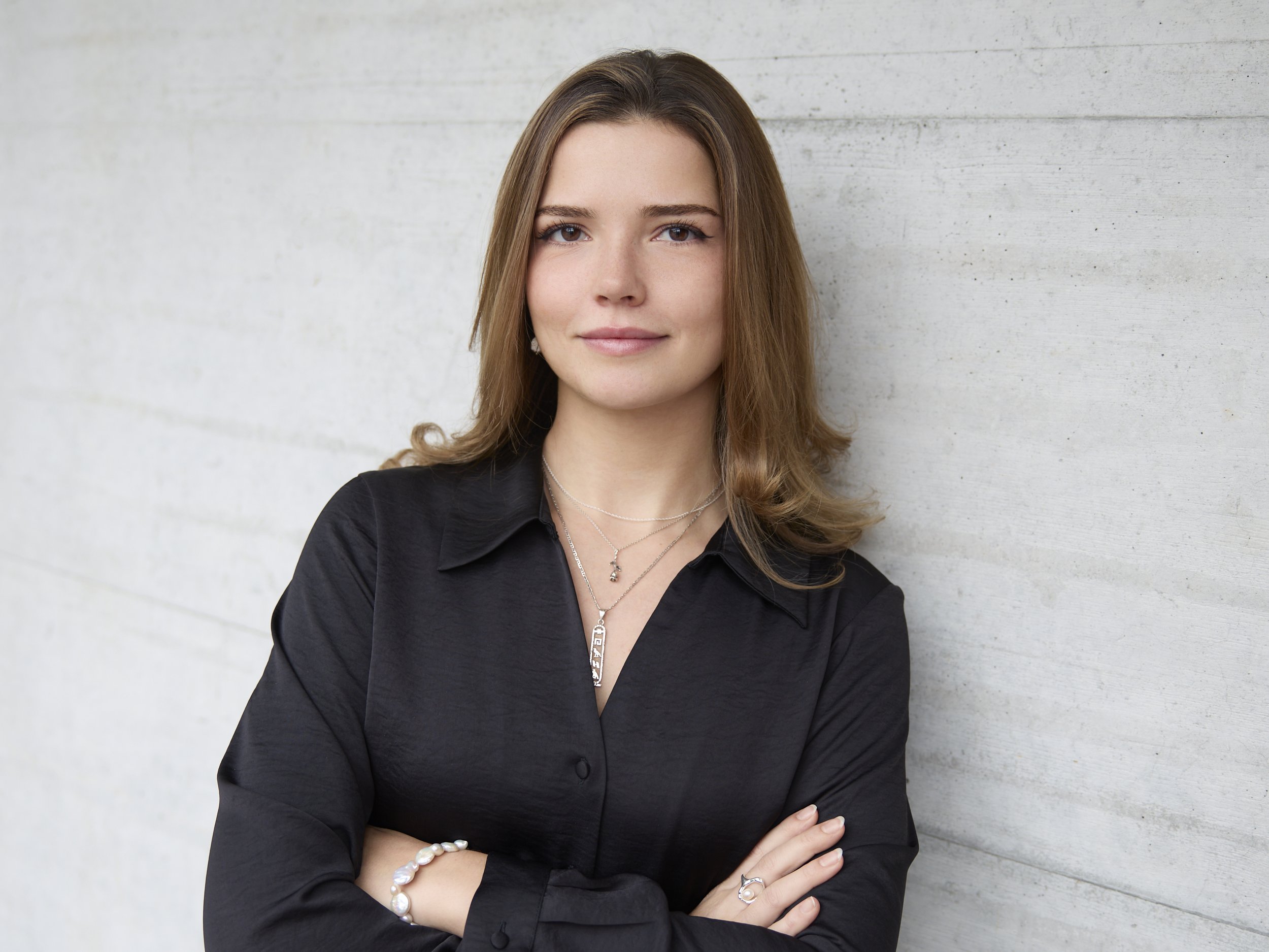 Junge Frau mit braunen Haaren und schwarzer Bluse, steht relaxt vor einer grauen Betonwand.