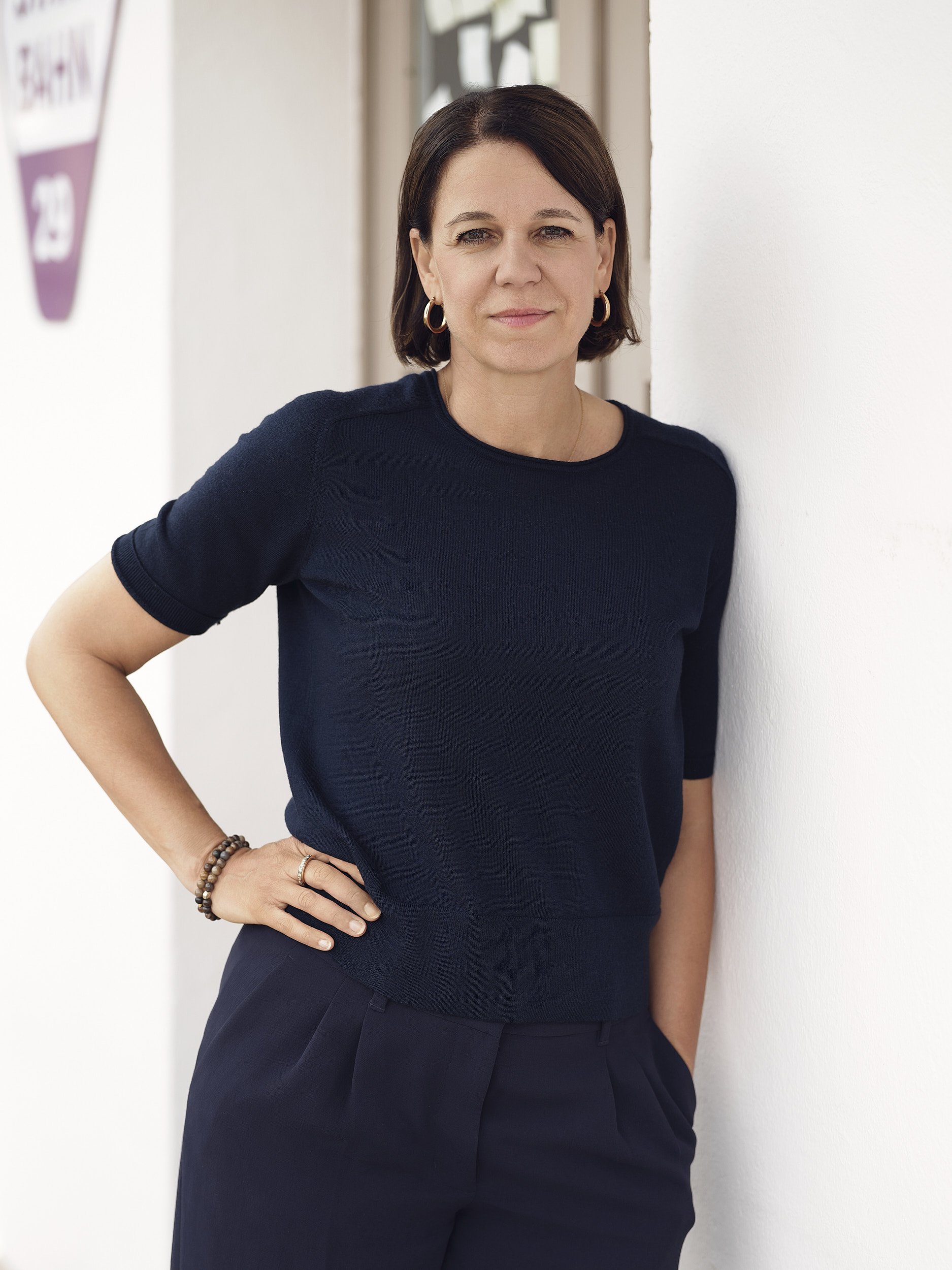 Portrait Fotografie. Eine Frau lehnt an einer weissen Wand und schaut direkt in die Kamera.