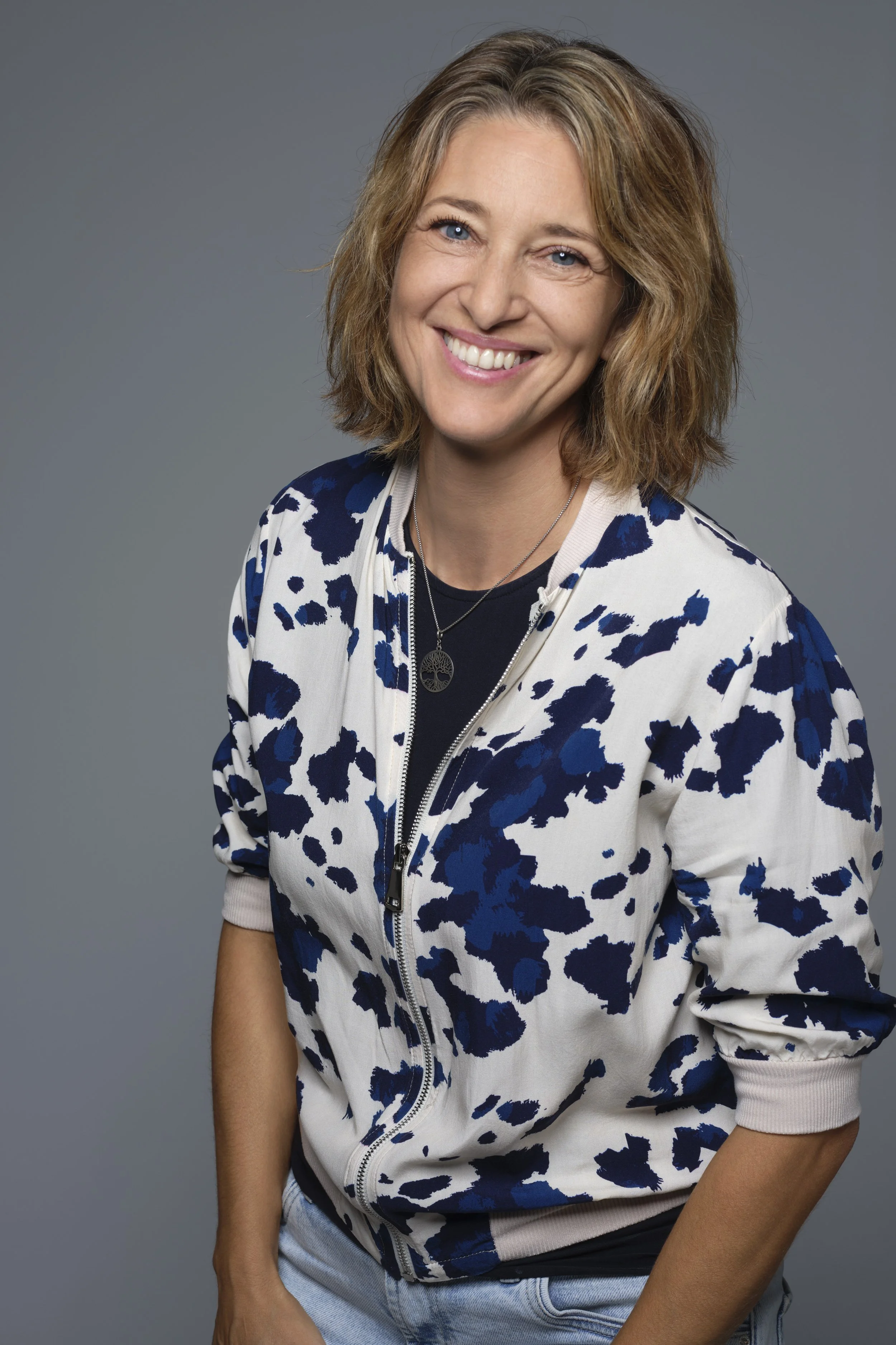 Eine lächelnde Frau mit braunen, lockigen Haaren in einem grauen Hintergrund, trägt eine weiße Jacke mit blauen Mustern.