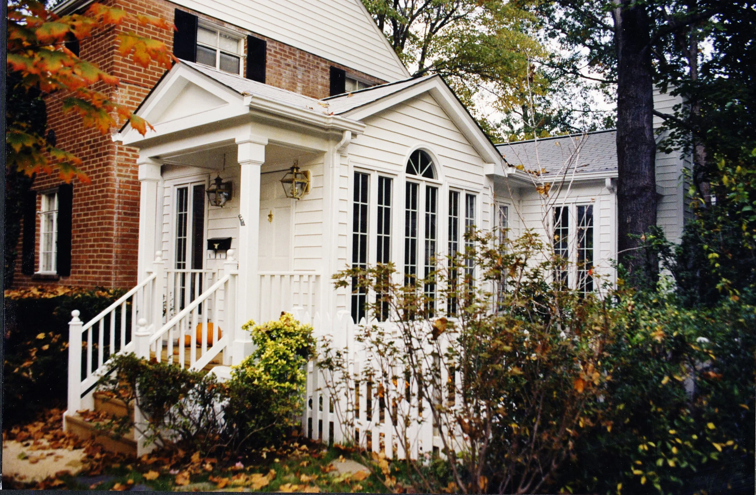 A small white house with a porch, surrounded by trees and bushes, with part of a brick building visible on the left.