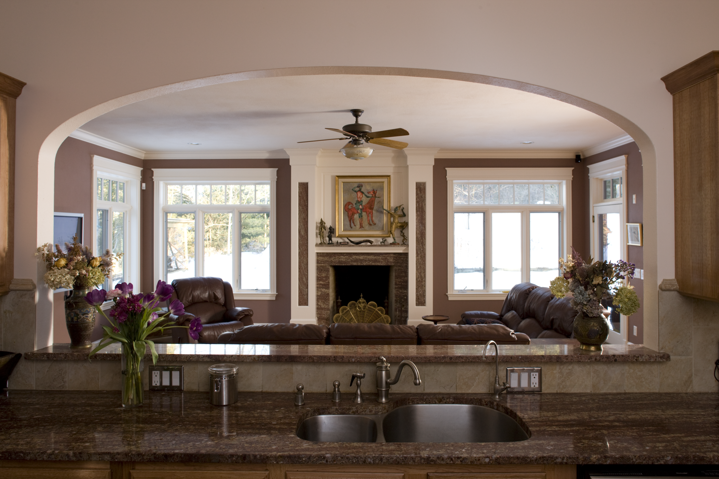 Living room with large windows, a fireplace, leather sofas, and floral arrangements on a kitchen counter.