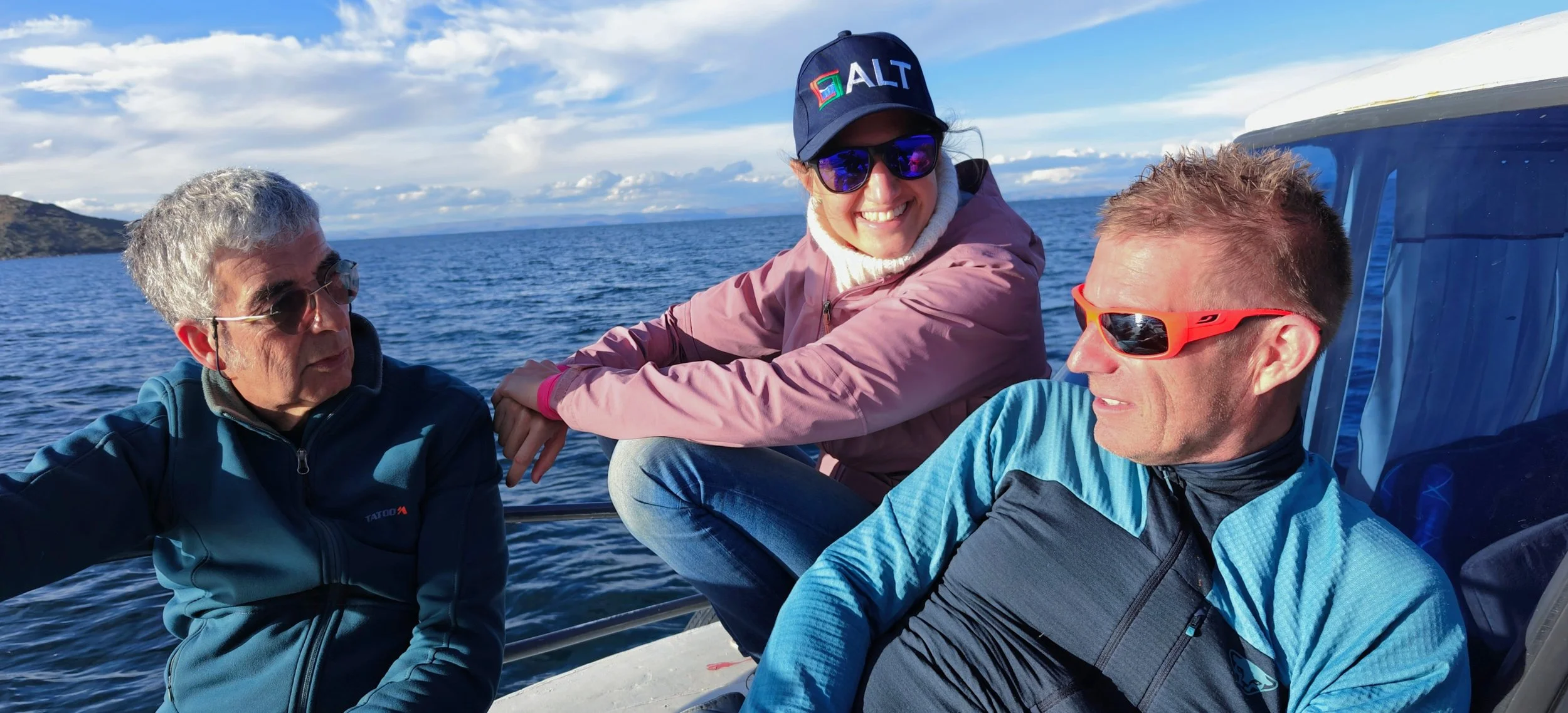 Three people sitting on a boat, enjoying a day on the water under a partly cloudy sky, with land visible in the background.