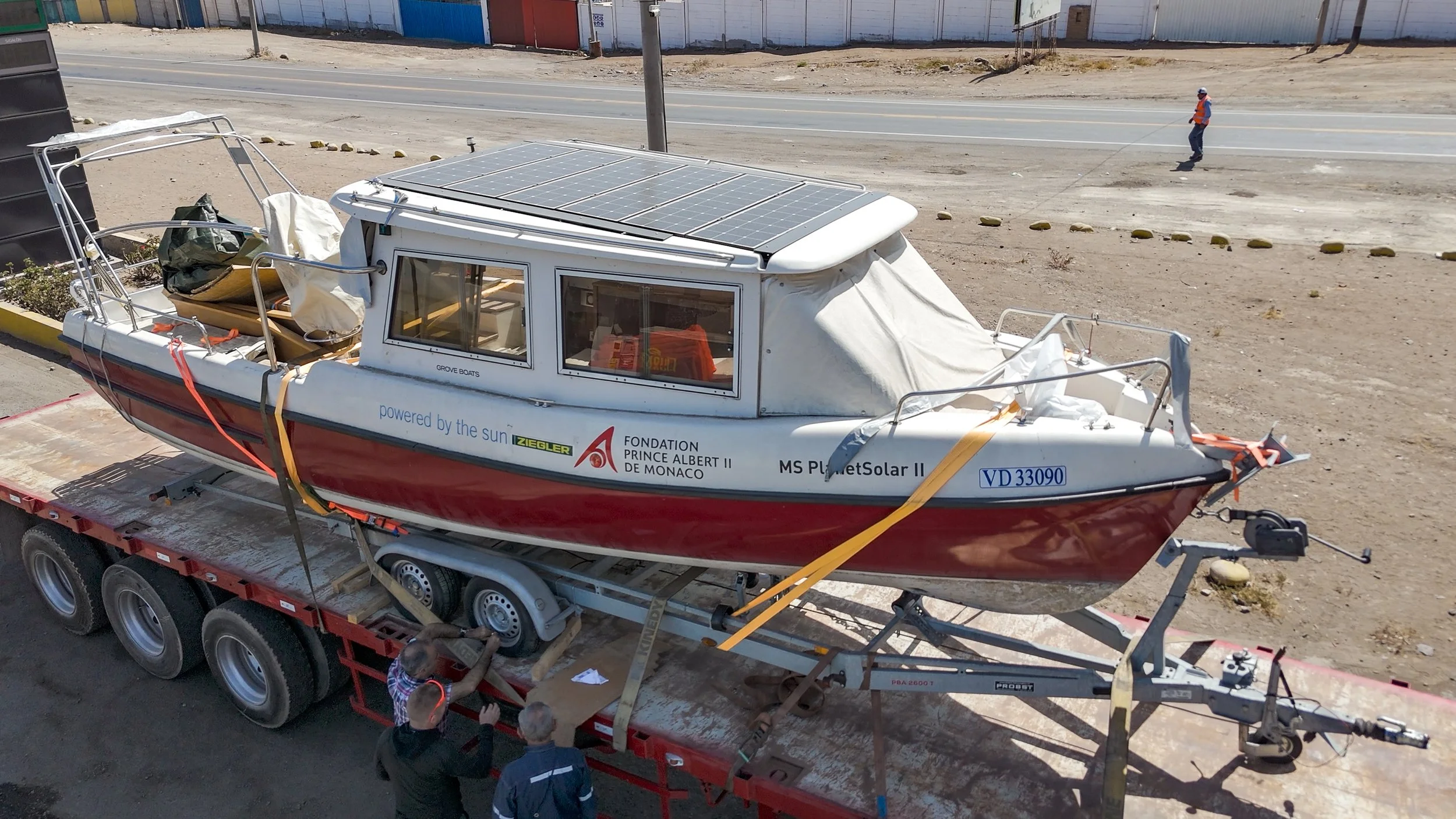 A small boat equipped with solar panels on top, mounted on a truck trailer in a desert-like area with a few people observing it.