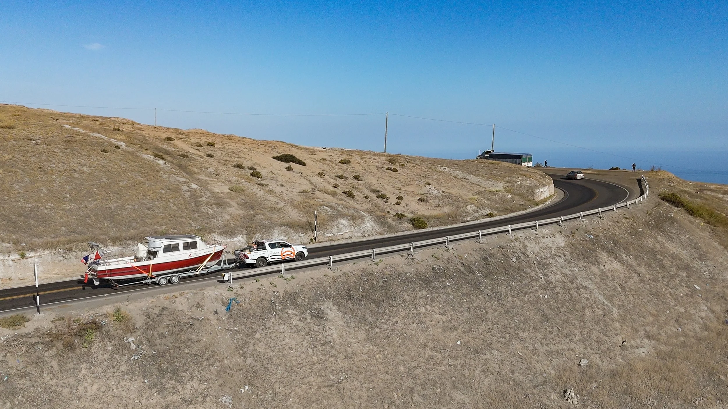 A winding coastal road on a hillside with a boat on a trailer being pulled by a police vehicle. The road has guardrails and there are a few people and vehicles in the distance, with the ocean visible in the background.
