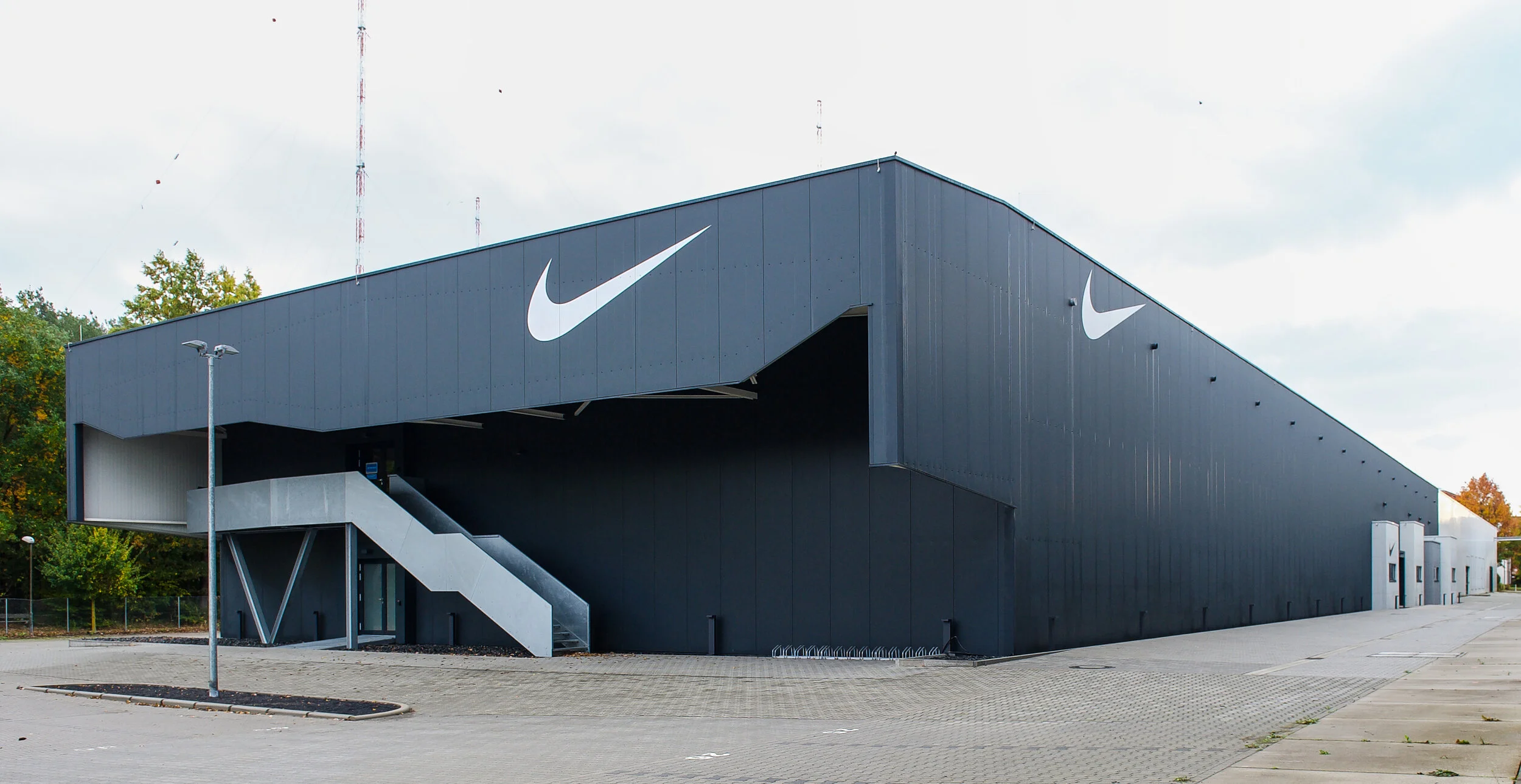 Schwarzes Gebäude mit großen weißen Nike-Logos und externen Treppenaufgänge, umgeben von einer gepflasterten Fläche und einigen Bäumen im Hintergrund.