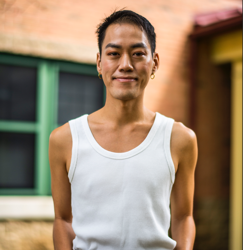 Portrait of a smiling person wearing a white tank top and gold hoop earrings, standing outdoors with a colorful brick wall and windows in the background.