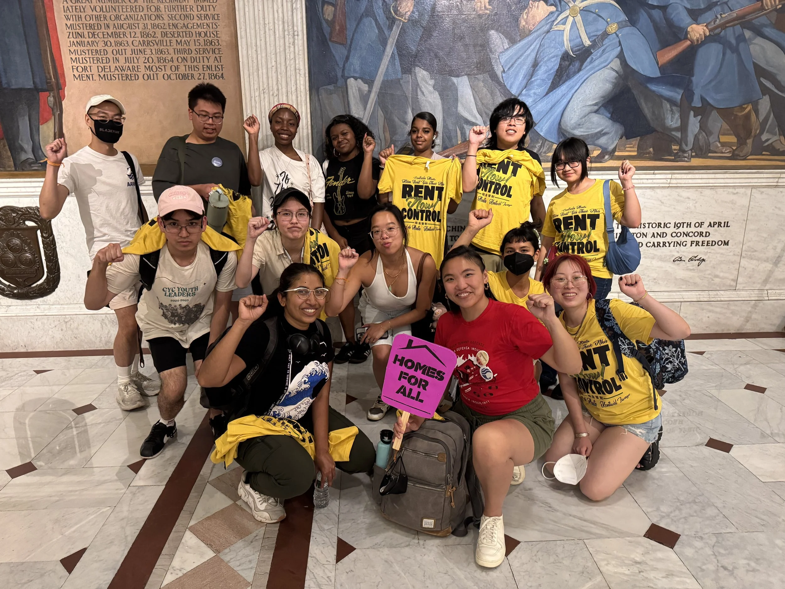 A group of young people, mostly women, posing with raised fists inside a historic building, some wearing yellow shirts with the words 'RENT CONTROL'. They are smiling, and one person is holding a pink sign that reads 'HOMES FOR ALL'.