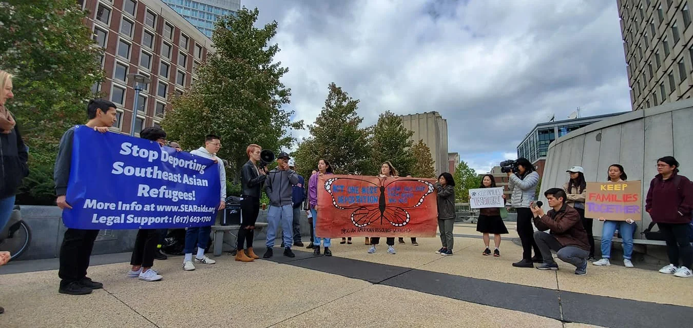 Protest in an urban area with people holding signs. One sign reads "Stop Deporting Southeast Asian Refugees!" Another sign says "Keep Families Together." A person is speaking into a megaphone, and several others are recording or taking pictures. The background features tall buildings, trees, and cloudy sky.