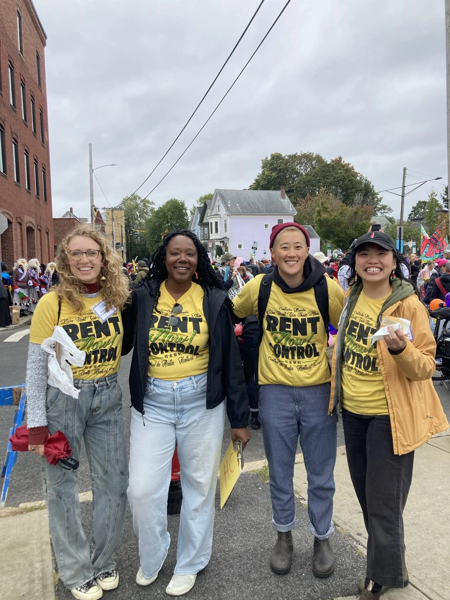 Four women wearing yellow T-shirts that promote rent control stand together on a city street during a parade or protest, smiling at the camera.