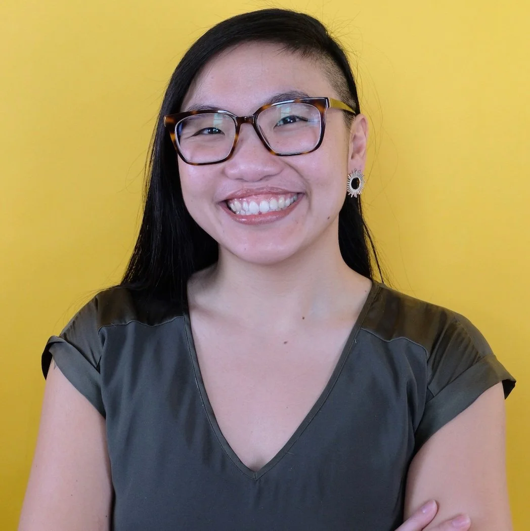 A young woman with long black hair, wearing glasses, earrings, and a dark green top, smiling in front of a yellow background.