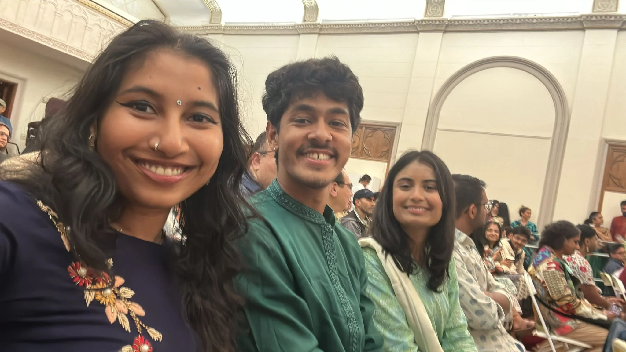 Three smiling young adults sitting in an audience at an indoor event, with people seated behind them near a large arched window.