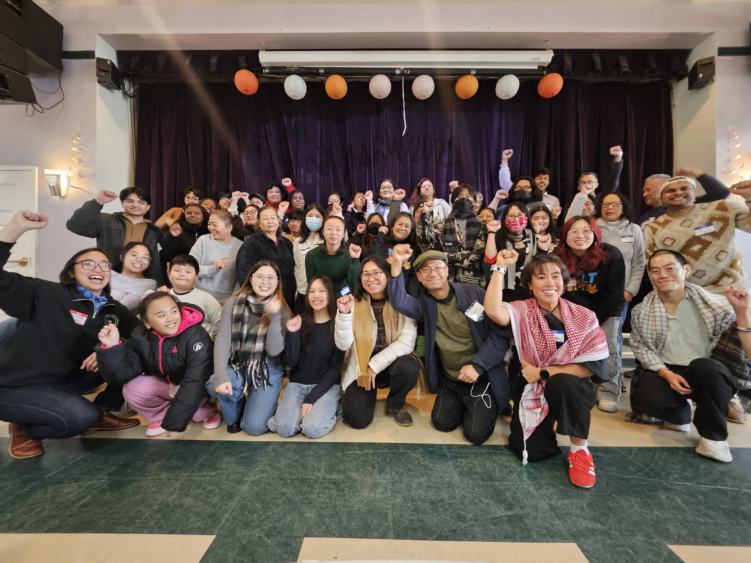 Group of diverse people posing together on a stage, smiling and raising fists, celebrating an event, with purple curtains and hanging paper lanterns in the background.