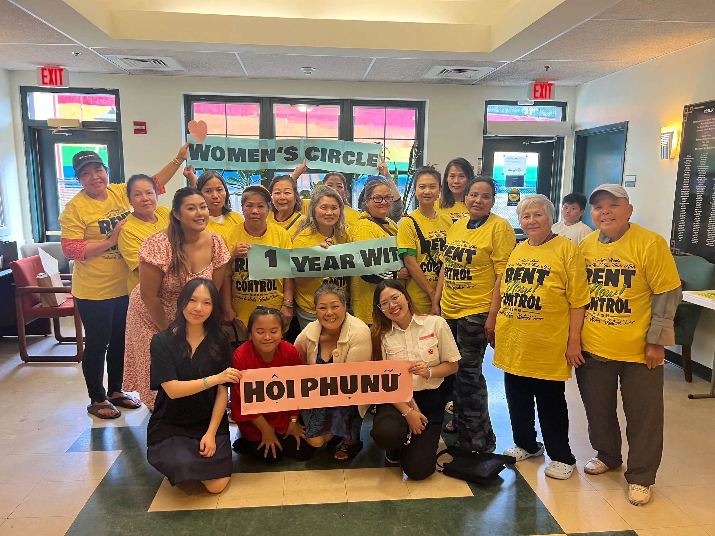 Group of women and girls standing inside a building holding a sign that says 'Women’s Circle 1 Year With HỘI PHỤ NỮ'. The women are wearing yellow shirts with black text, some are smiling, and there are windows and exit signs in the background.