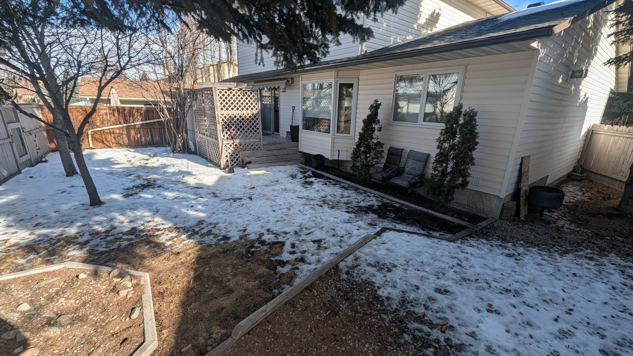 Backyard scene with snow on the ground, a tree on the left, a wooden deck with lattice fencing, and outdoor chairs near the house.