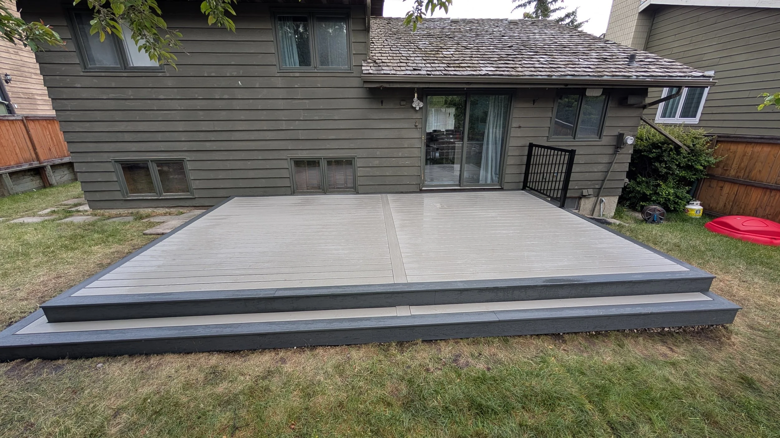 Backyard with a new raised deck in front of a brown house with gray siding, sliding glass door, and small windows, surrounded by a wooden fence and green grass.