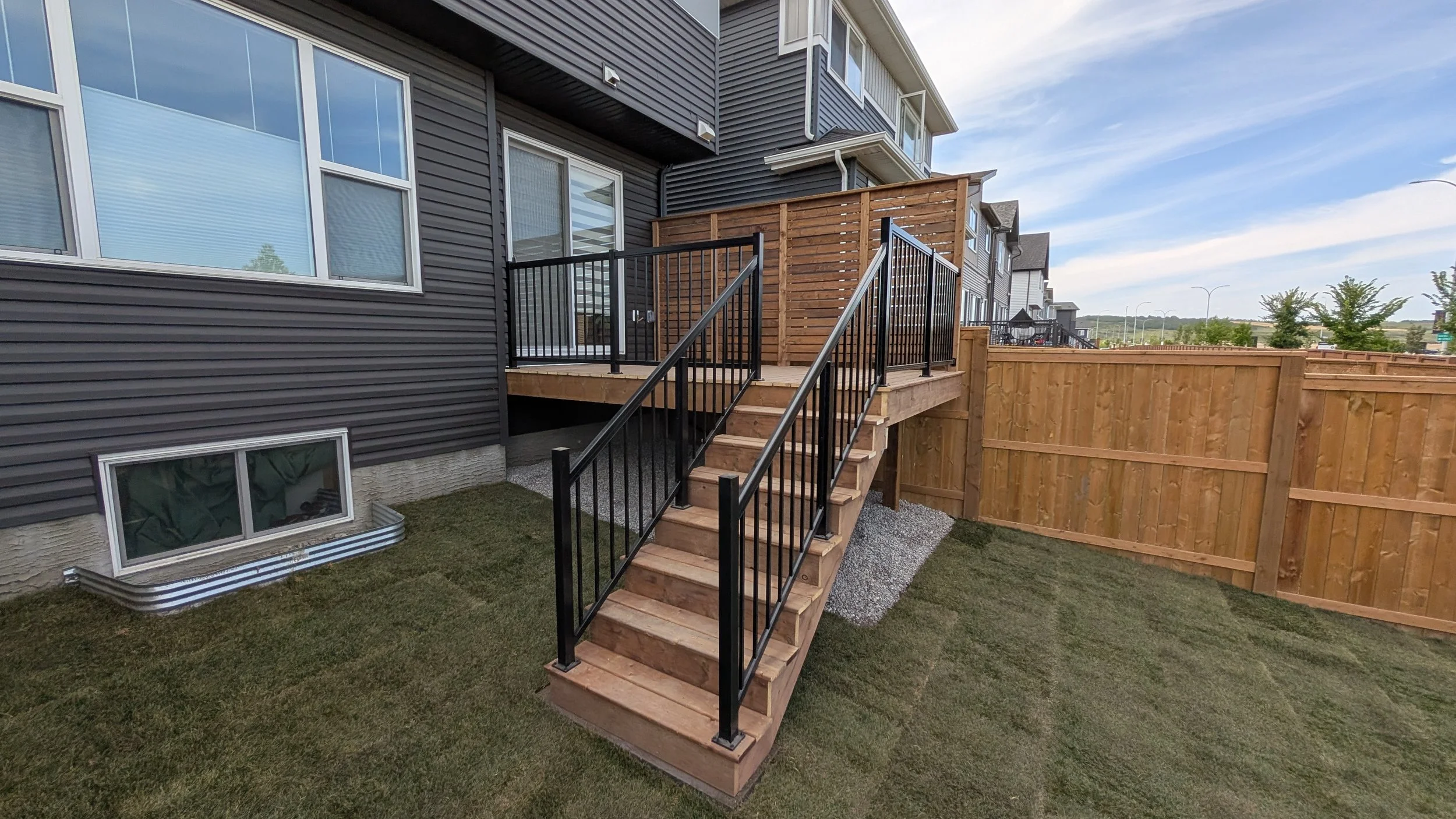 Newly built wooden outdoor staircase with metal railings leading up to a patio deck in a residential backyard, enclosed by a wooden fence.