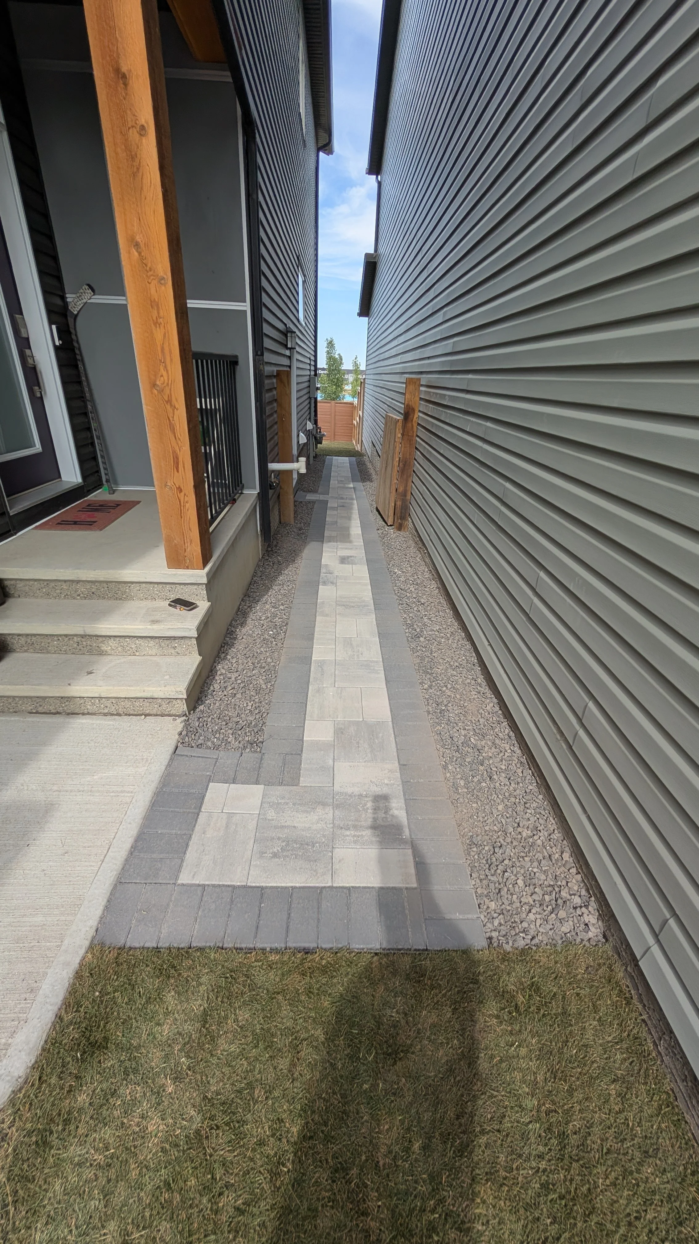A narrow pathway between two houses, with concrete steps leading up to a door on the left and a small grassy area in the foreground. The pathway is paved with rectangular grey and white tiles, and there are some wooden posts on the sides.
