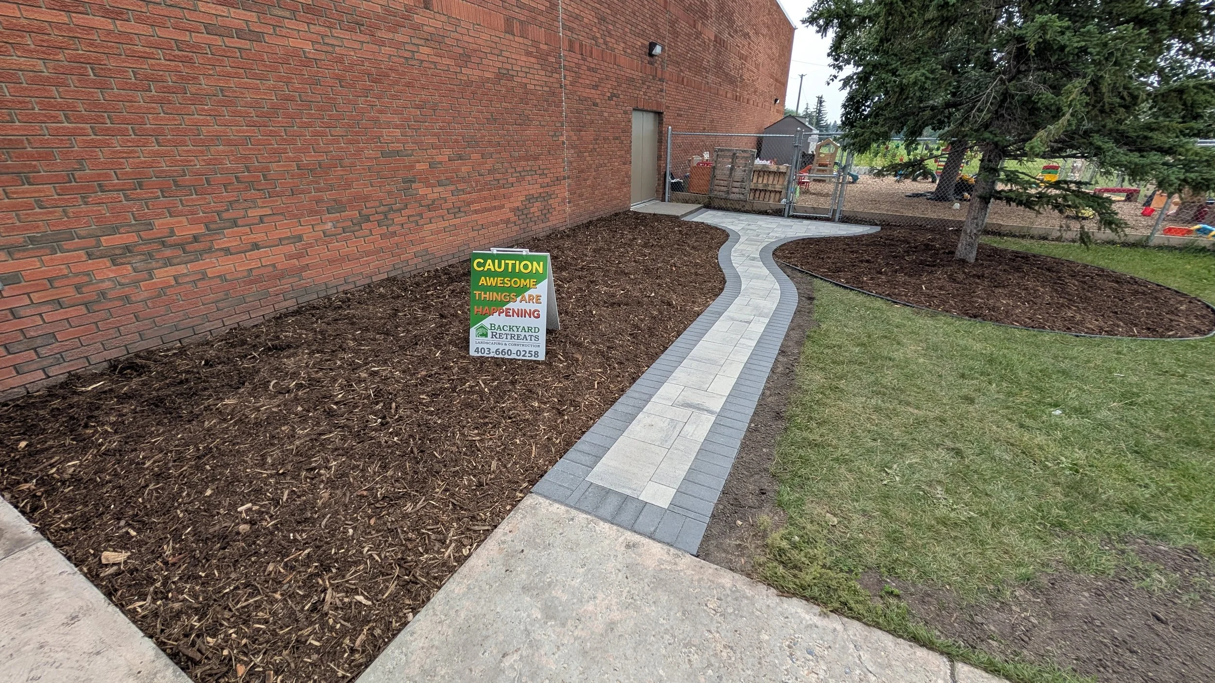 Newly paved sidewalk with a winding design alongside a brick building and a grassy area with a tree, with a sign that says 'CAUTION AWESOME THINGS ARE HAPPENING' near the sidewalk.