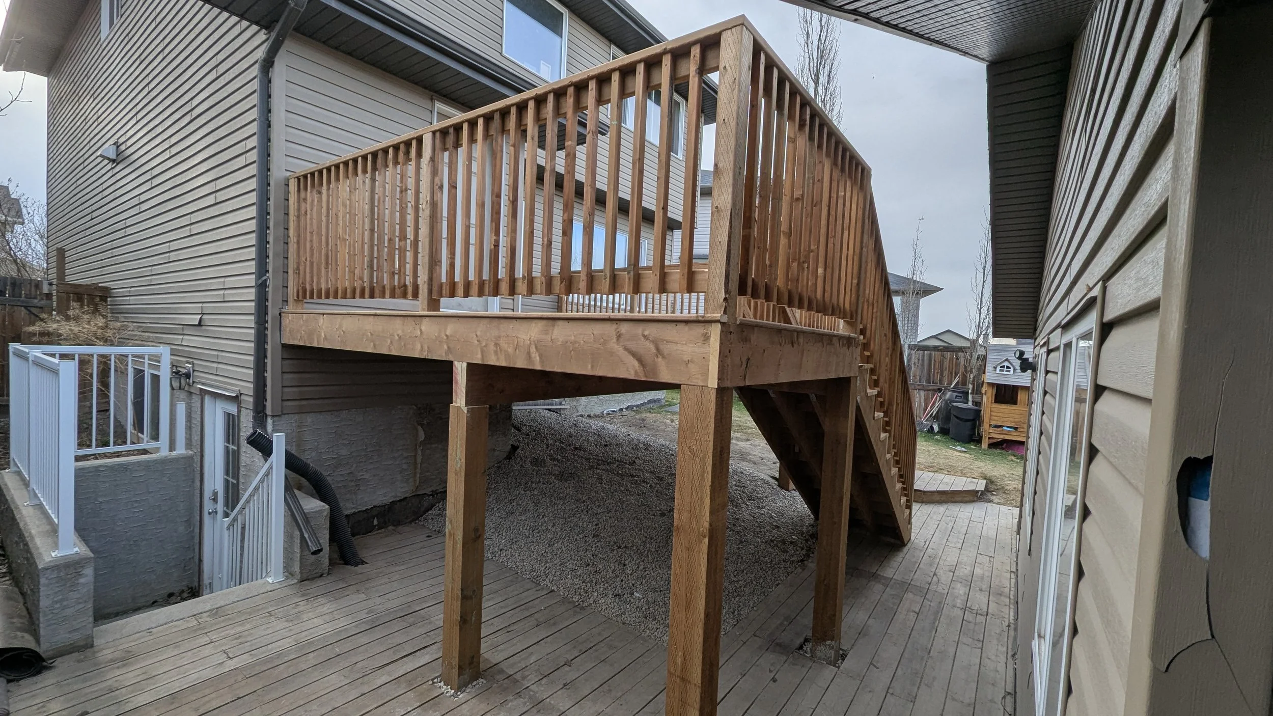 New wooden deck built with stairs and railing, elevated above a lower level, attached to a beige house with vinyl siding.