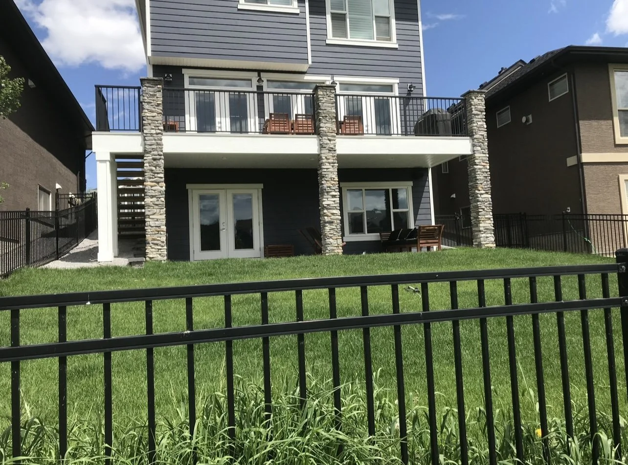 Modern multi-story house with a backyard, green lawn, stone pillars, and outdoor seating area, fenced with black metal fencing.