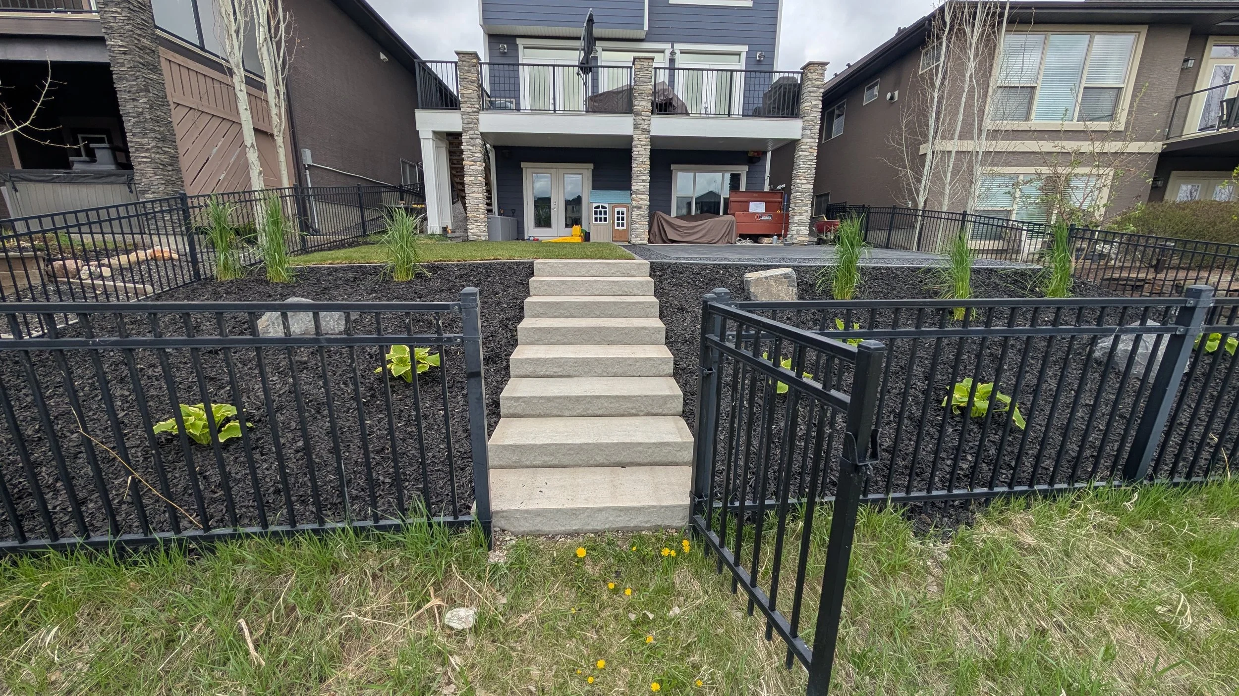 View of a backyard with a concrete staircase leading up to a house with a covered porch. The yard is landscaped with dark mulch, green plants, and bordered by a black metal fence. The house has a second-floor balcony, stone columns, and large windows