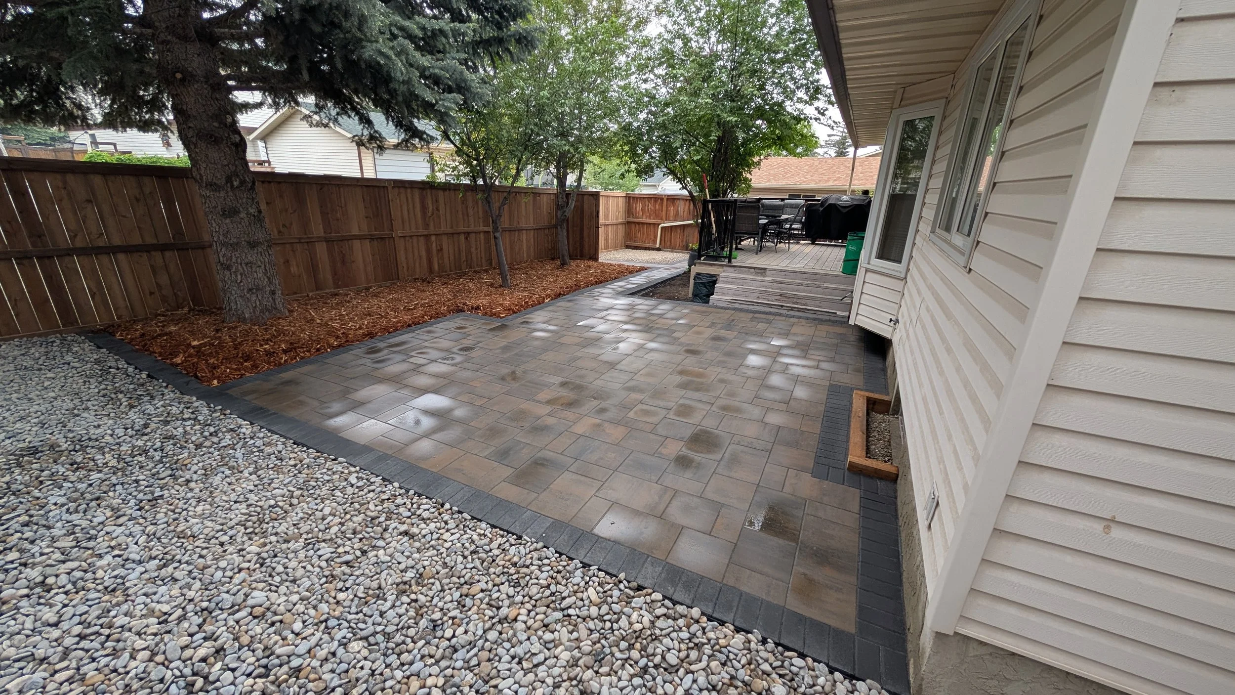 Backyard patio with stone and pebble surfaces, wooden deck with outdoor furniture, fenced yard with trees, and house siding.