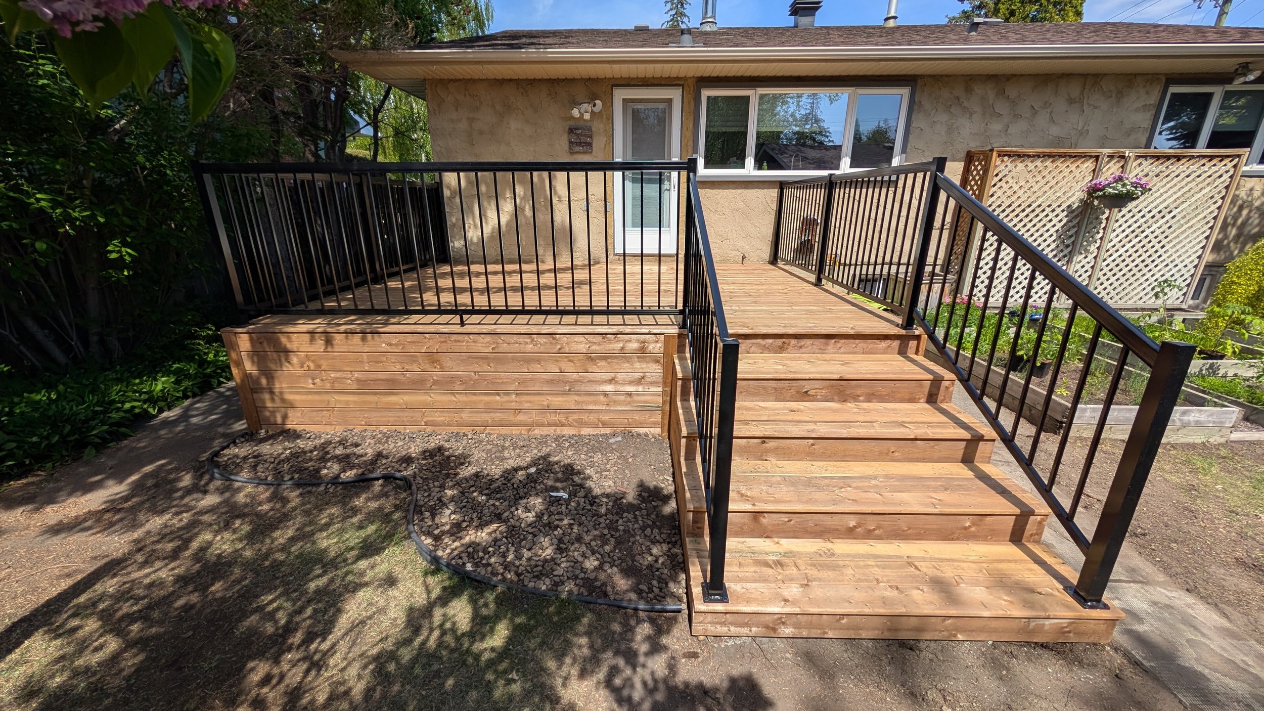 Newly built wooden deck with black metal railings attached to the back of a house, surrounded by trees and some garden beds.