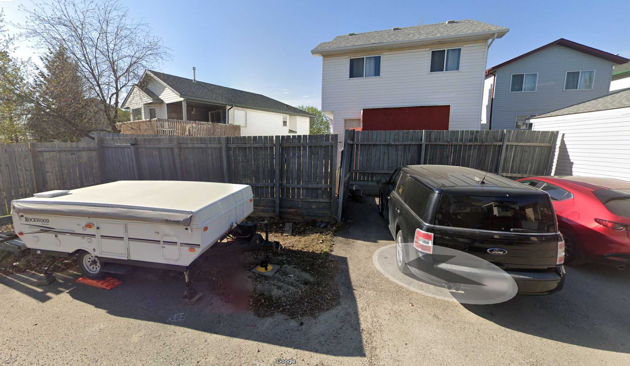 A parking lot with a white travel trailer on the left, a black SUV in the middle, and a red car on the right, all parked in front of a wooden fence and residential buildings.