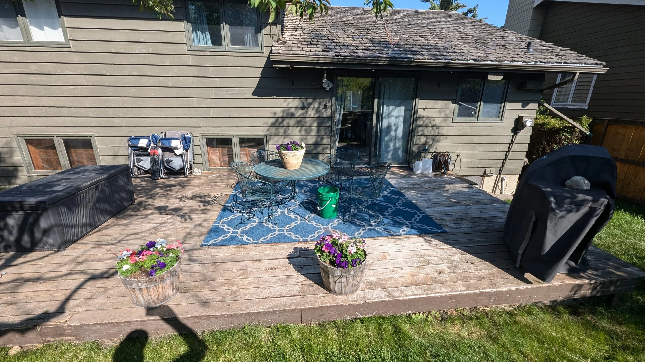 Backyard deck with patio furniture, potted flowers, a rug, a barbecue grill, and gardening supplies, with the house's exterior wall and windows in the background.