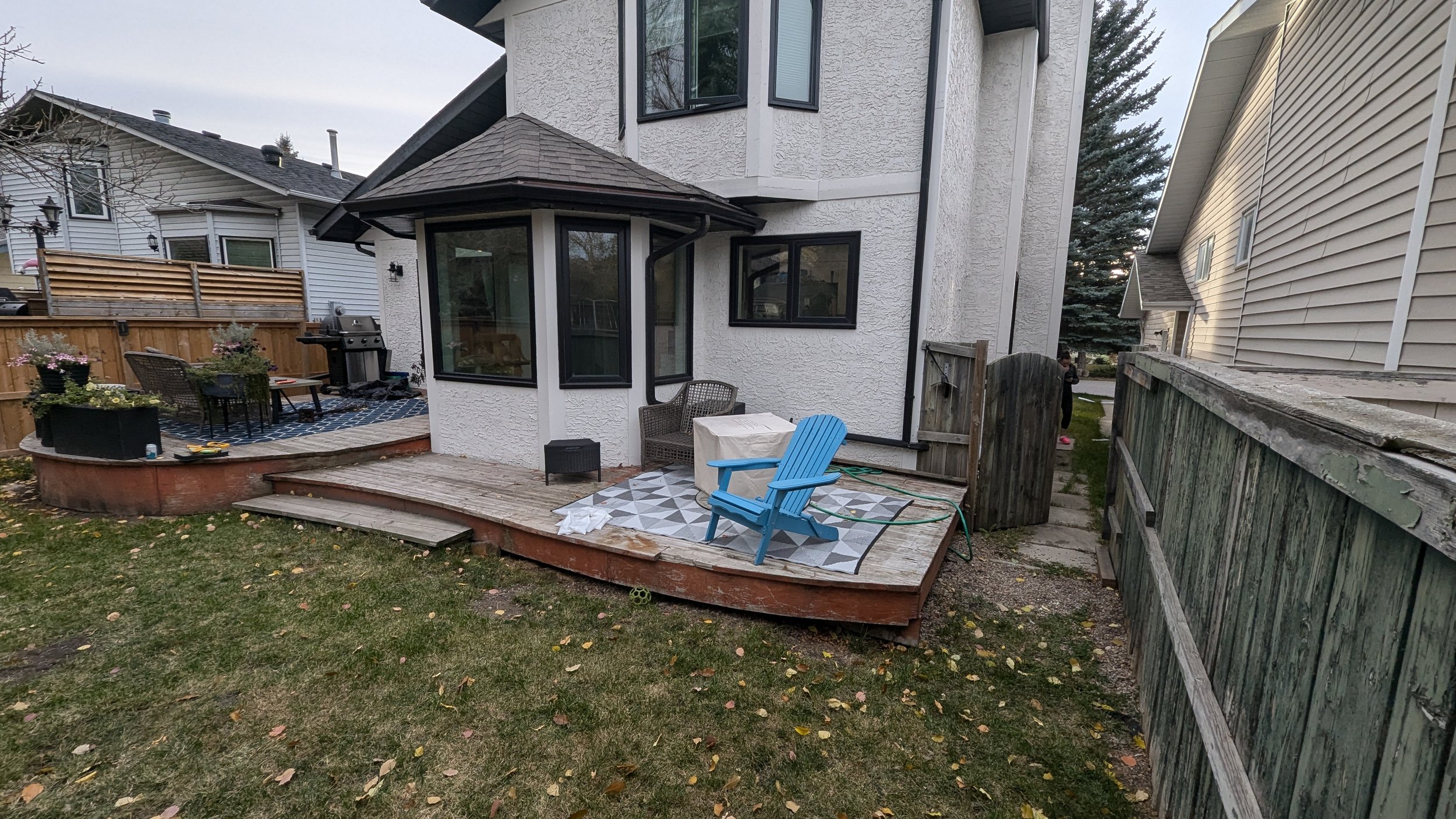 Backyard patio with a small wooden deck, a blue Adirondack chair, a patterned outdoor rug, and a covered table with a tablecloth. There are potted plants and a barbecue grill on the patio, and a neighboring house is visible in the background.