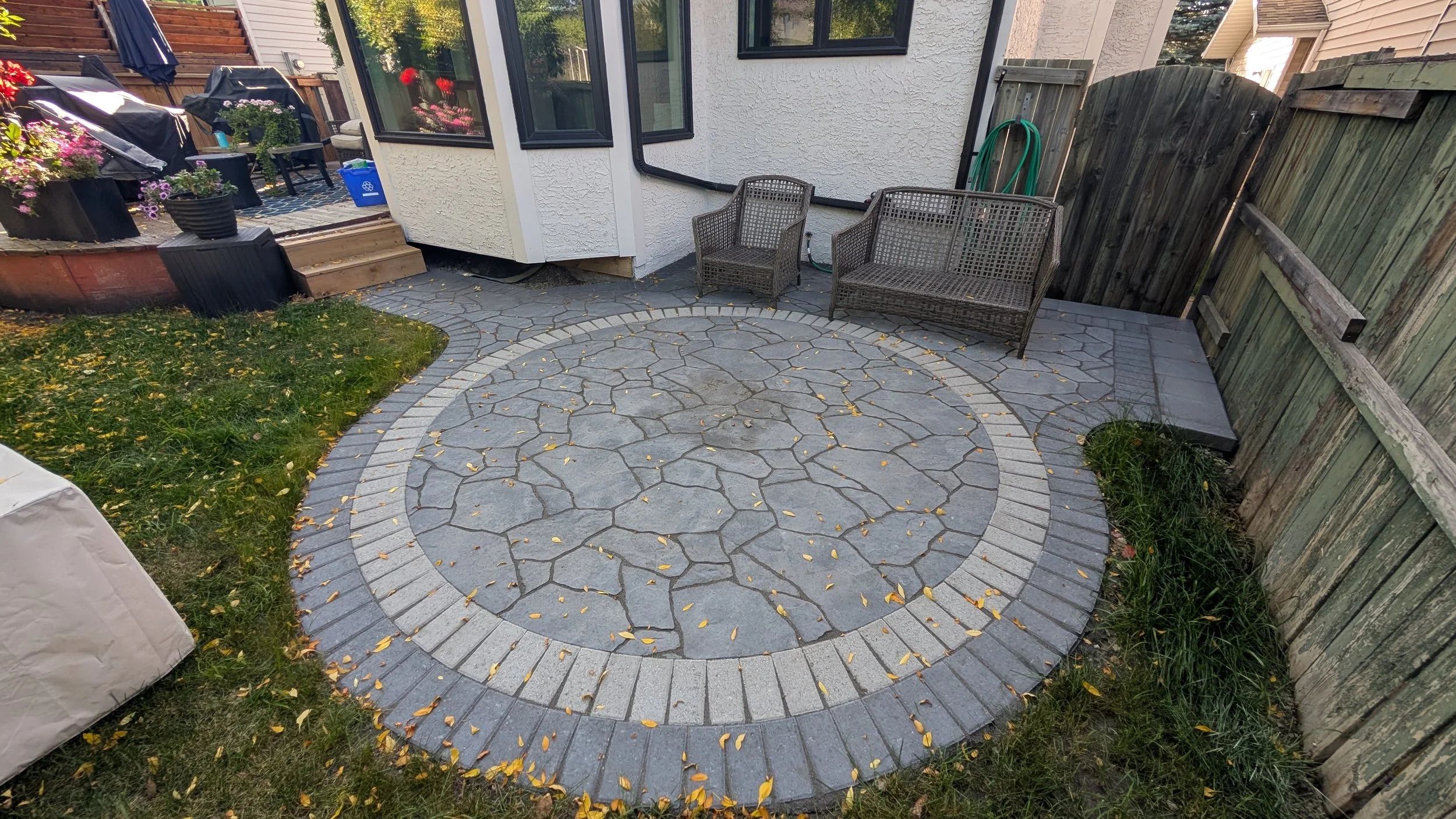 A backyard patio with circular stone paving, two wicker chairs, a small wicker table, and a garden hose near a white house with black windows and a wooden fence.