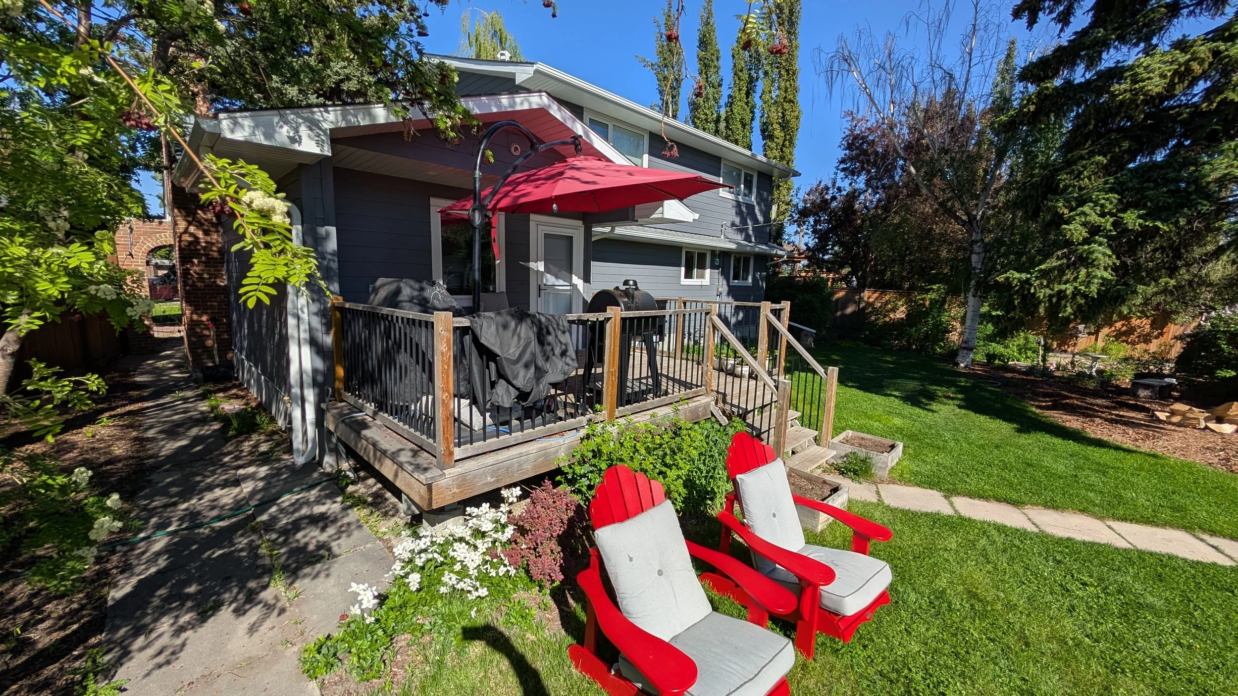Backyard with a wooden deck, red patio umbrella, grill, two red and white outdoor chairs, green grass, trees, and a blue sky.