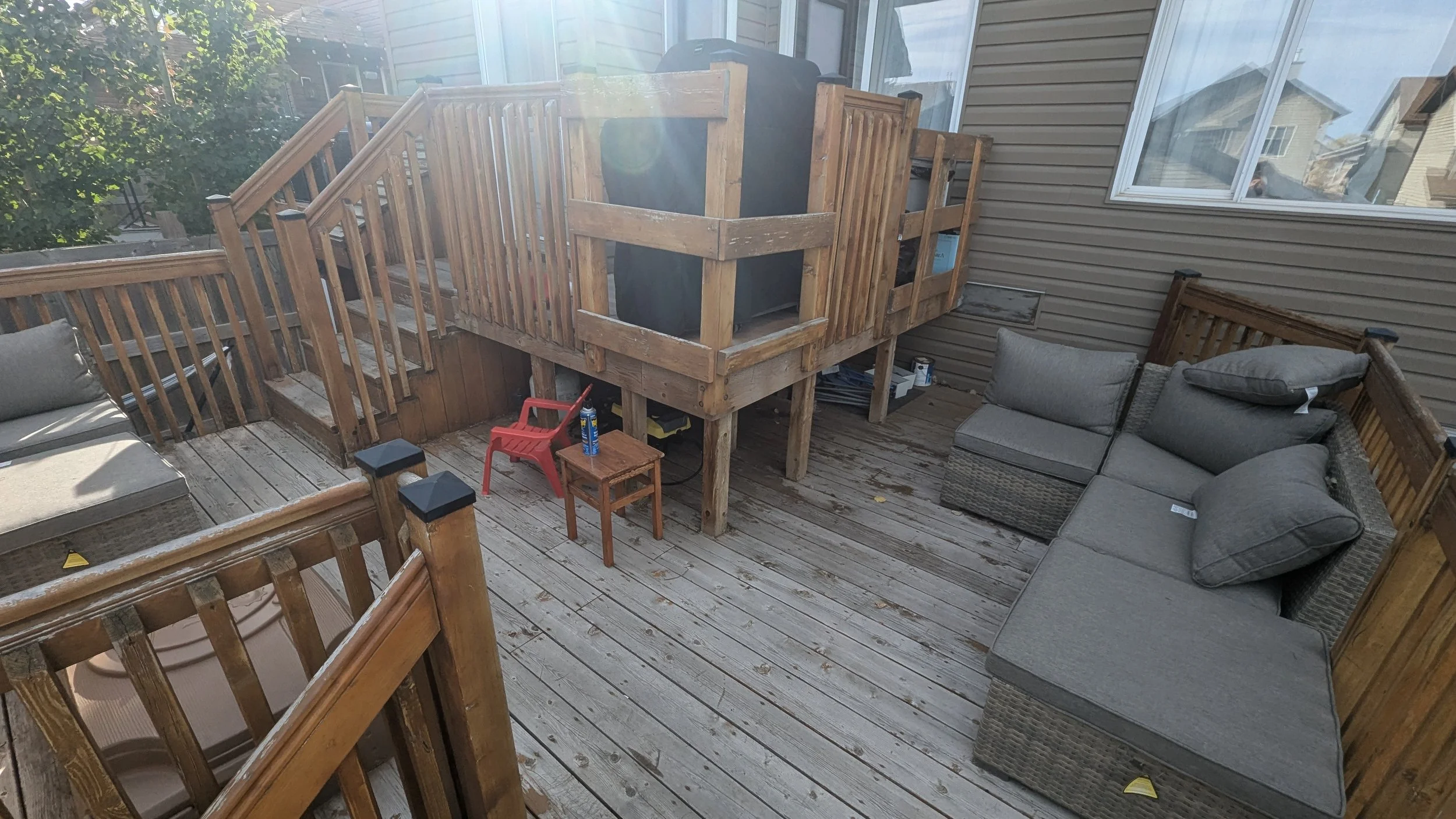A wooden outdoor deck with seating and a raised deck area with stairs, surrounded by a railing, attached to a house with beige siding and a large window, with a small red plastic chair and a small wooden table on the deck.