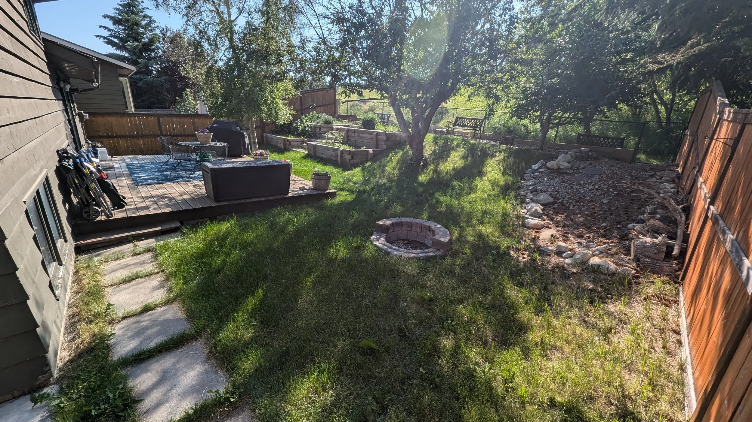 Backyard with wooden deck, lawn, fire pit, garden beds, trees, and a fence.