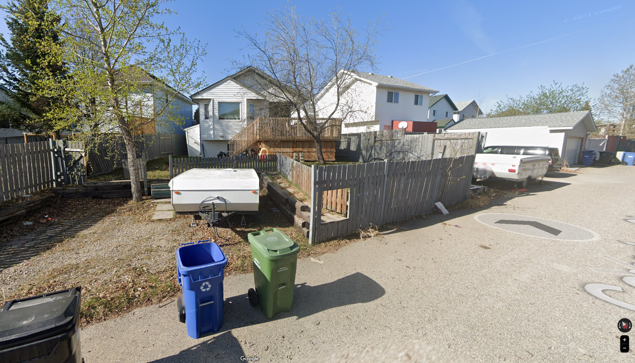 Residential backyard with a tree, a small fenced yard, a camper, trash bins, and neighboring houses, under a clear blue sky.