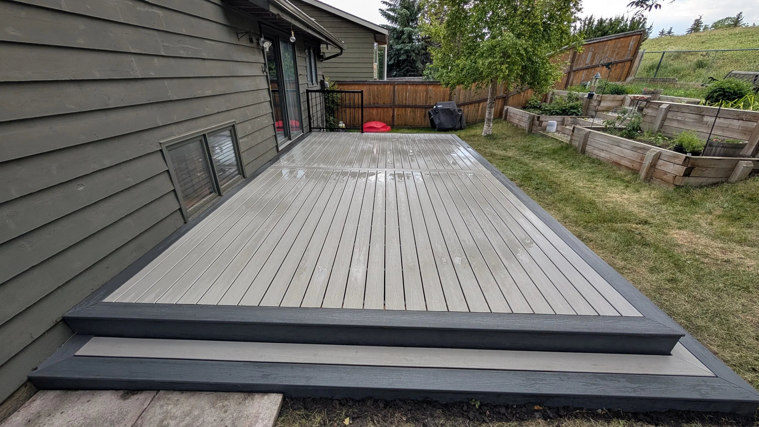 Backyard deck with wet surface next to house with multiple small windows, green siding, and outdoor string lights, with a lawn, garden beds, and a tree in the background.