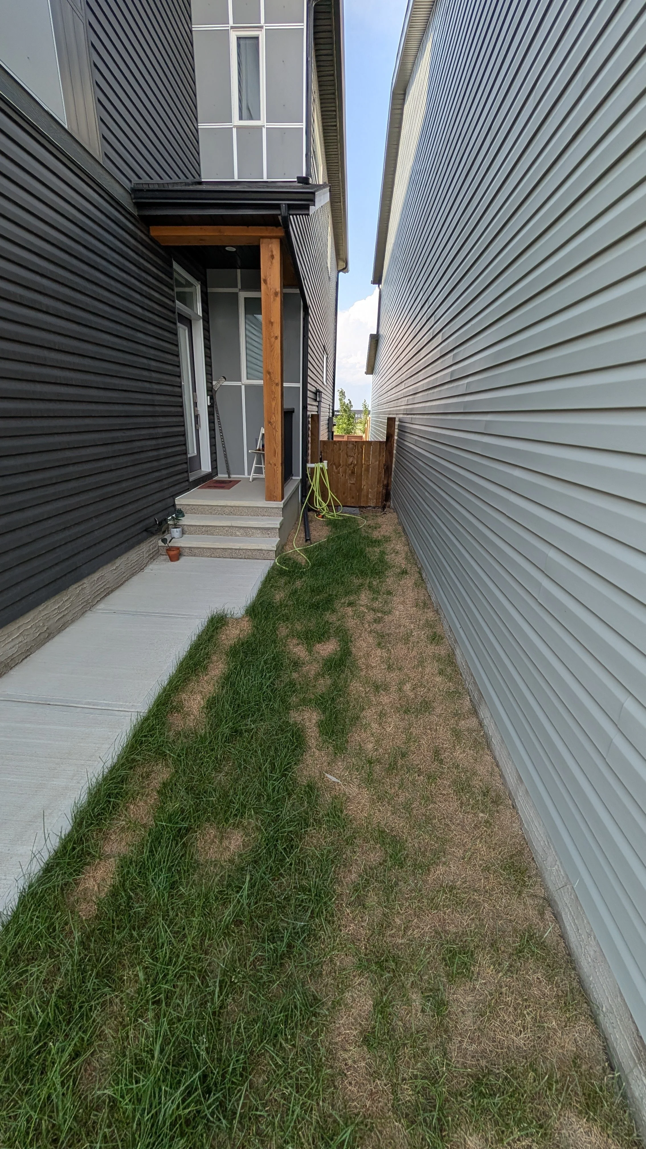 Narrow backyard between two houses with siding, a small concrete patio, stairs leading to a door, a hose, and a small fenced area at the end.