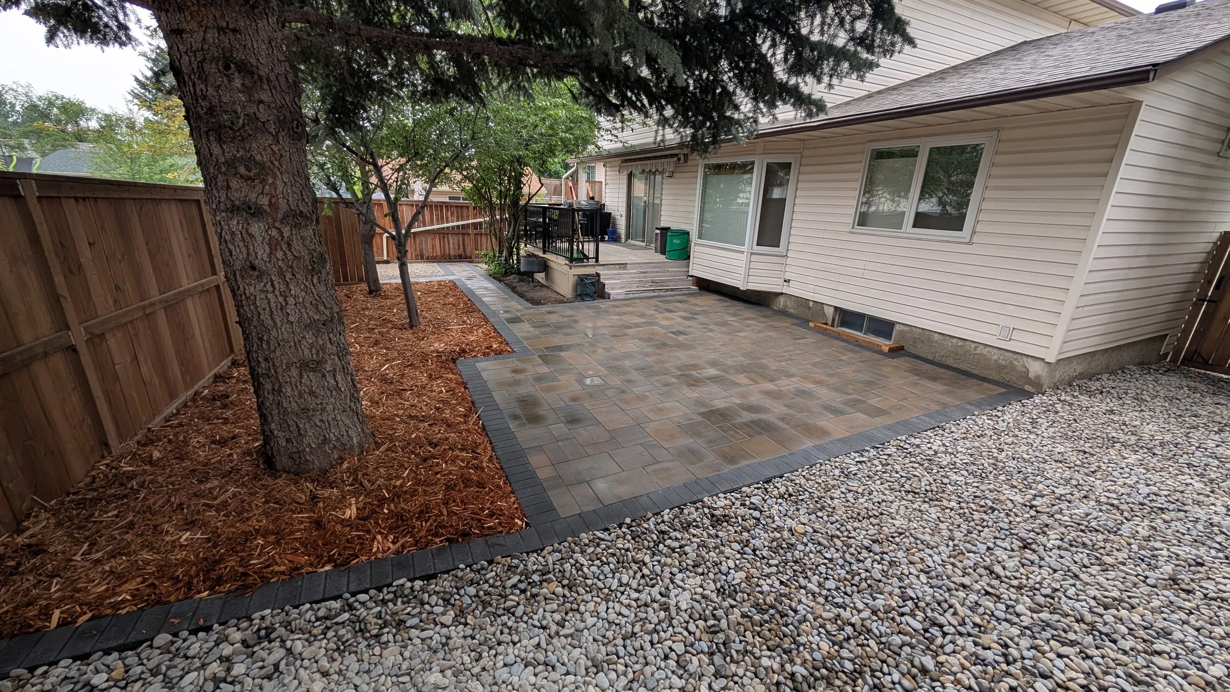 Backyard with a large tree, mulch bed, stone and brick patio, and a sliding glass door leading to the house.